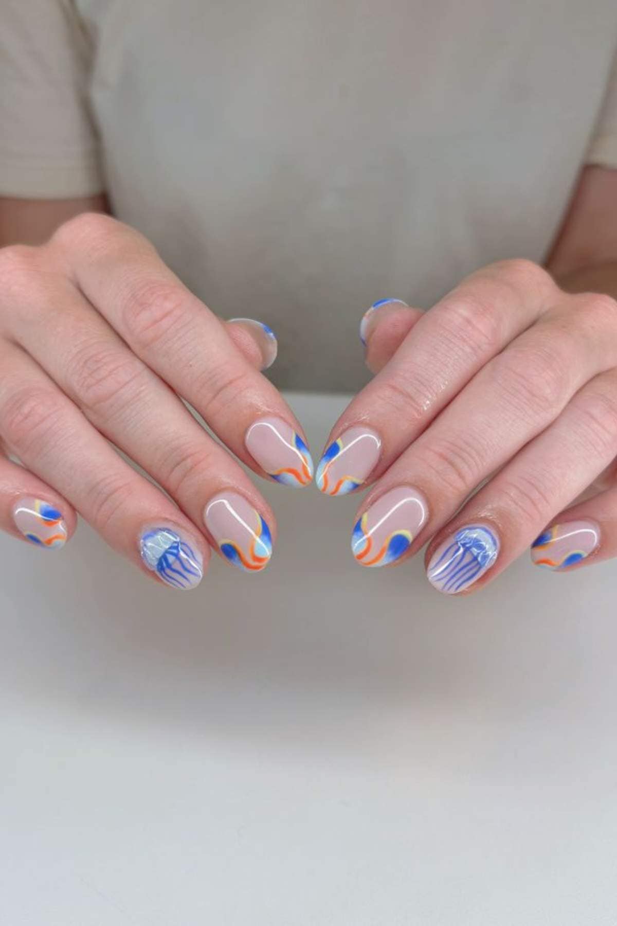 A person with short, rounded nails featuring abstract blue, orange, and white designs, including swirls and lines on a neutral base.