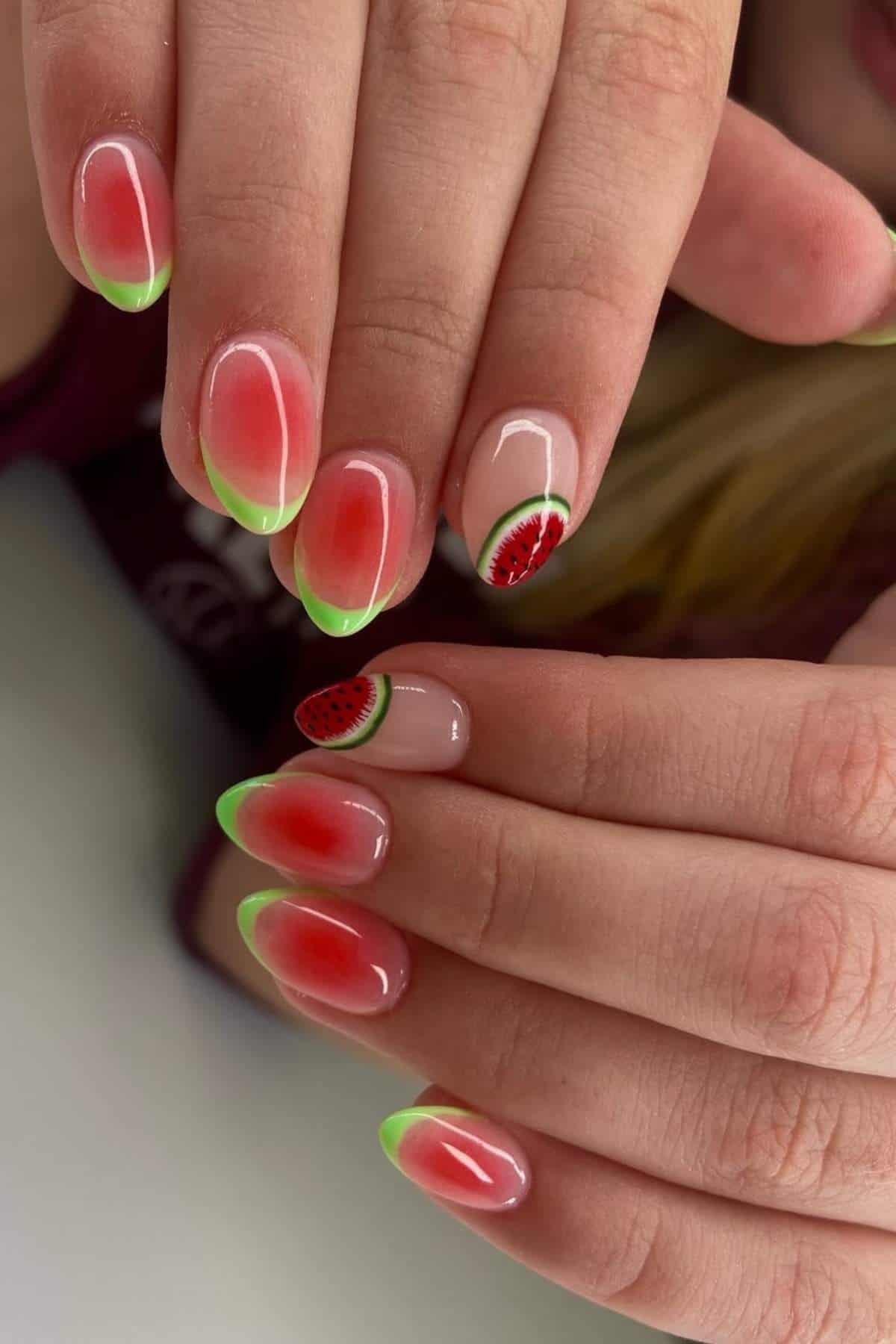 Hands with manicure featuring red and green ombre nails; two nails have watermelon slice designs near the cuticles.