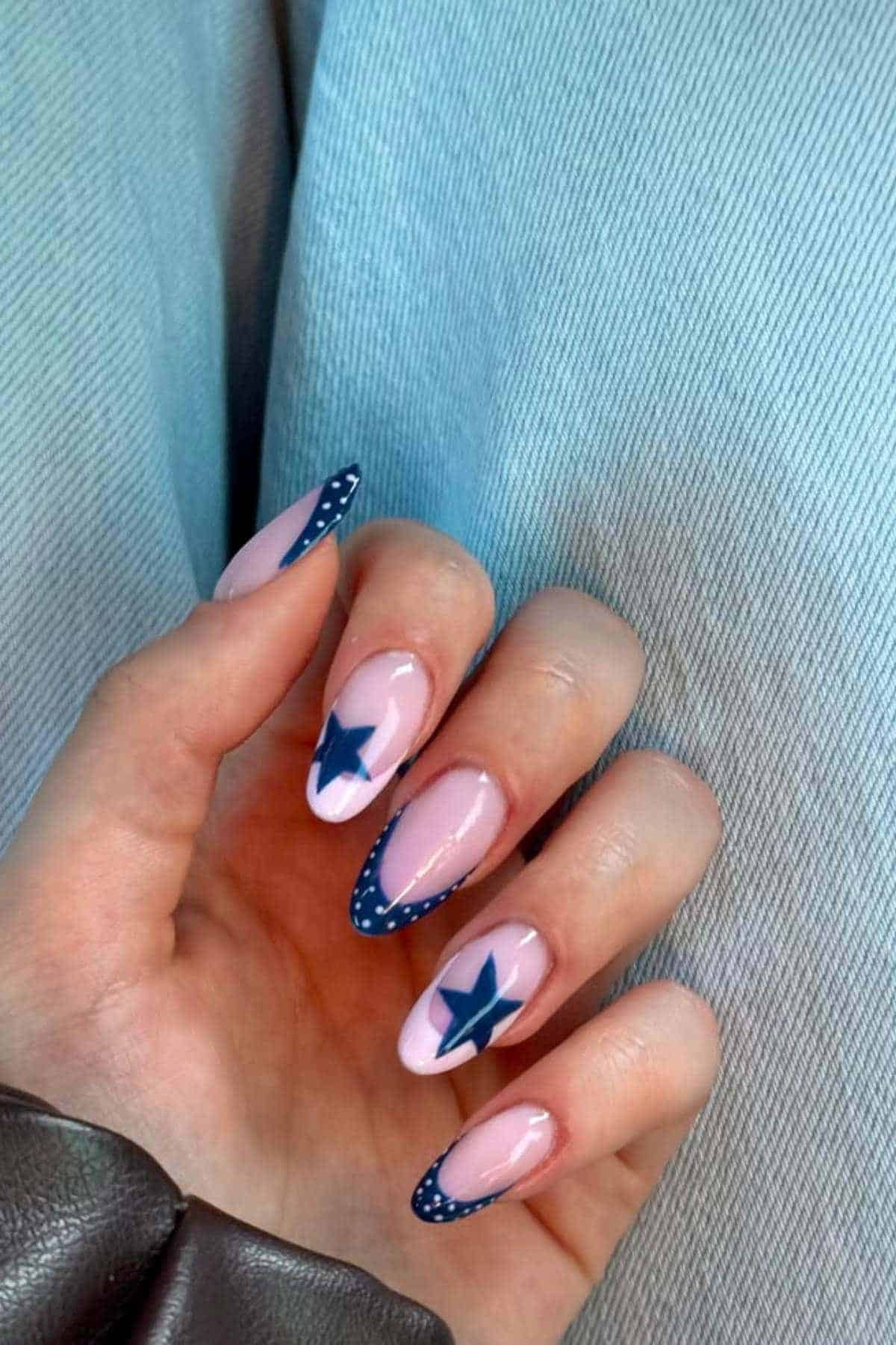 A hand with long, almond-shaped nails painted light pink, featuring navy blue star designs and dotted navy blue French tips, resting on light blue denim fabric.