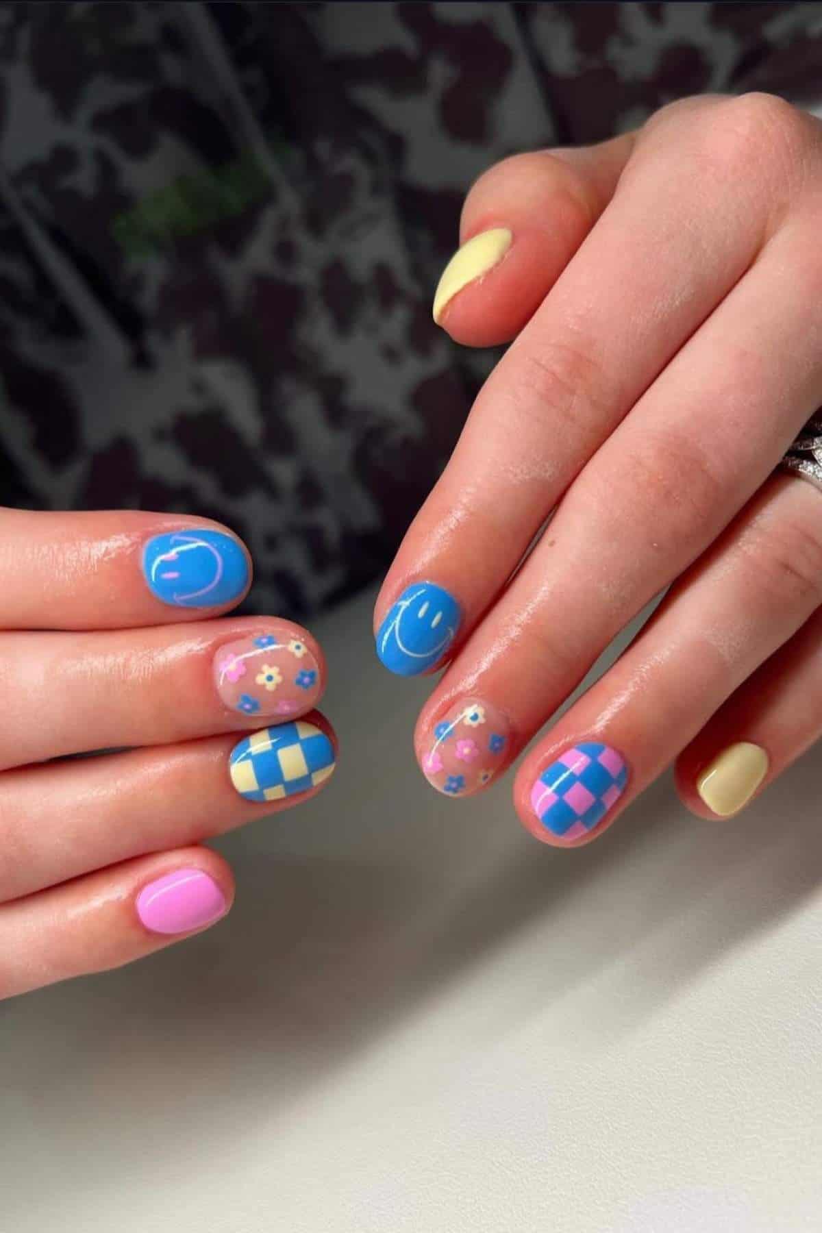Hands with multicolored nail art featuring pastel blue, pink, and yellow, with designs including flowers, smiley faces, and checkerboard patterns.