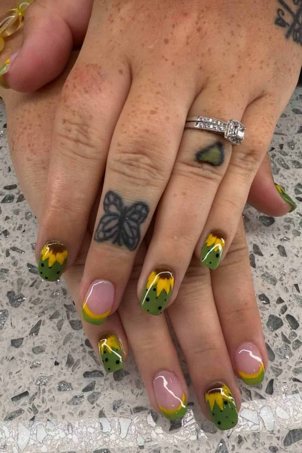 Hands with green and yellow sunflower-themed nail art, butterfly and heart tattoos, and a diamond ring on a speckled countertop.