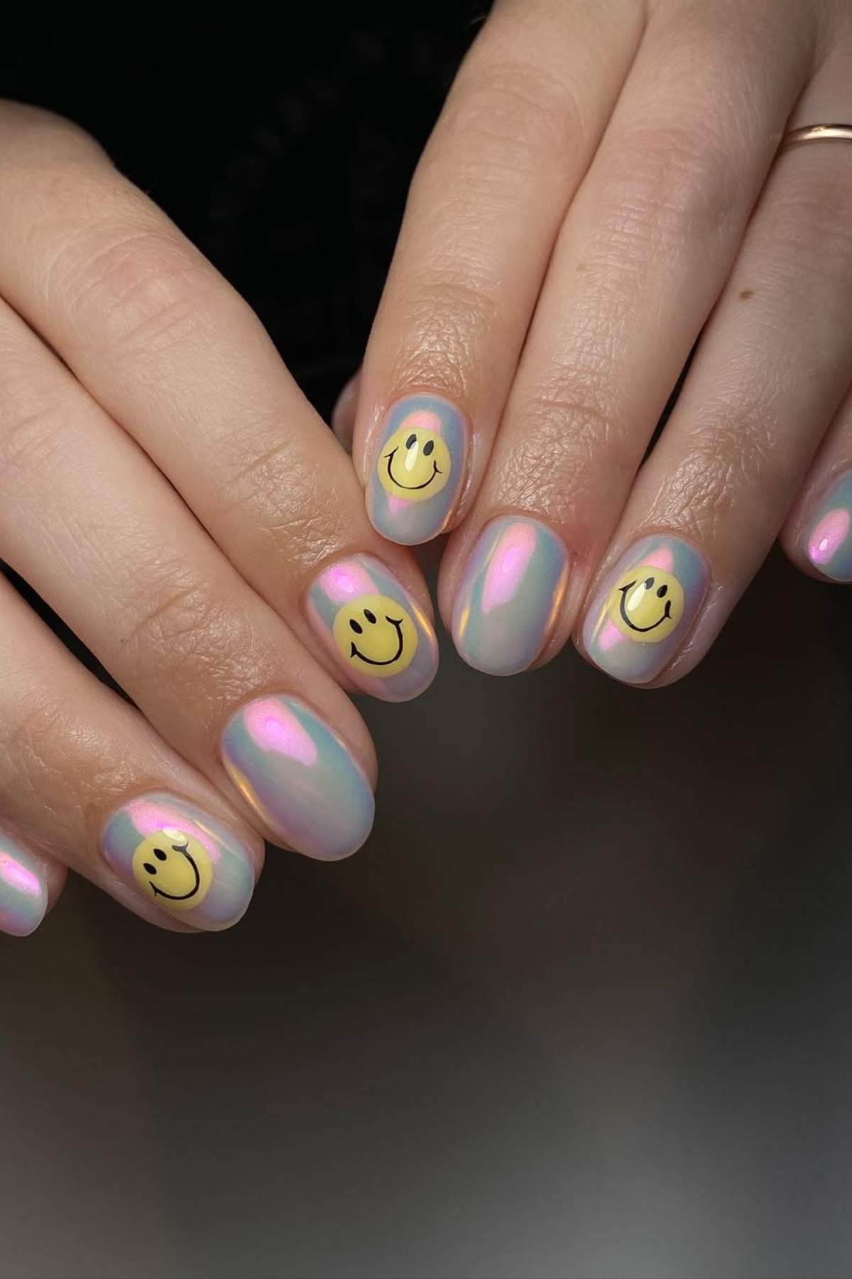 Hands with iridescent pink nails, each featuring a yellow smiley face design on the middle and ring fingernails.