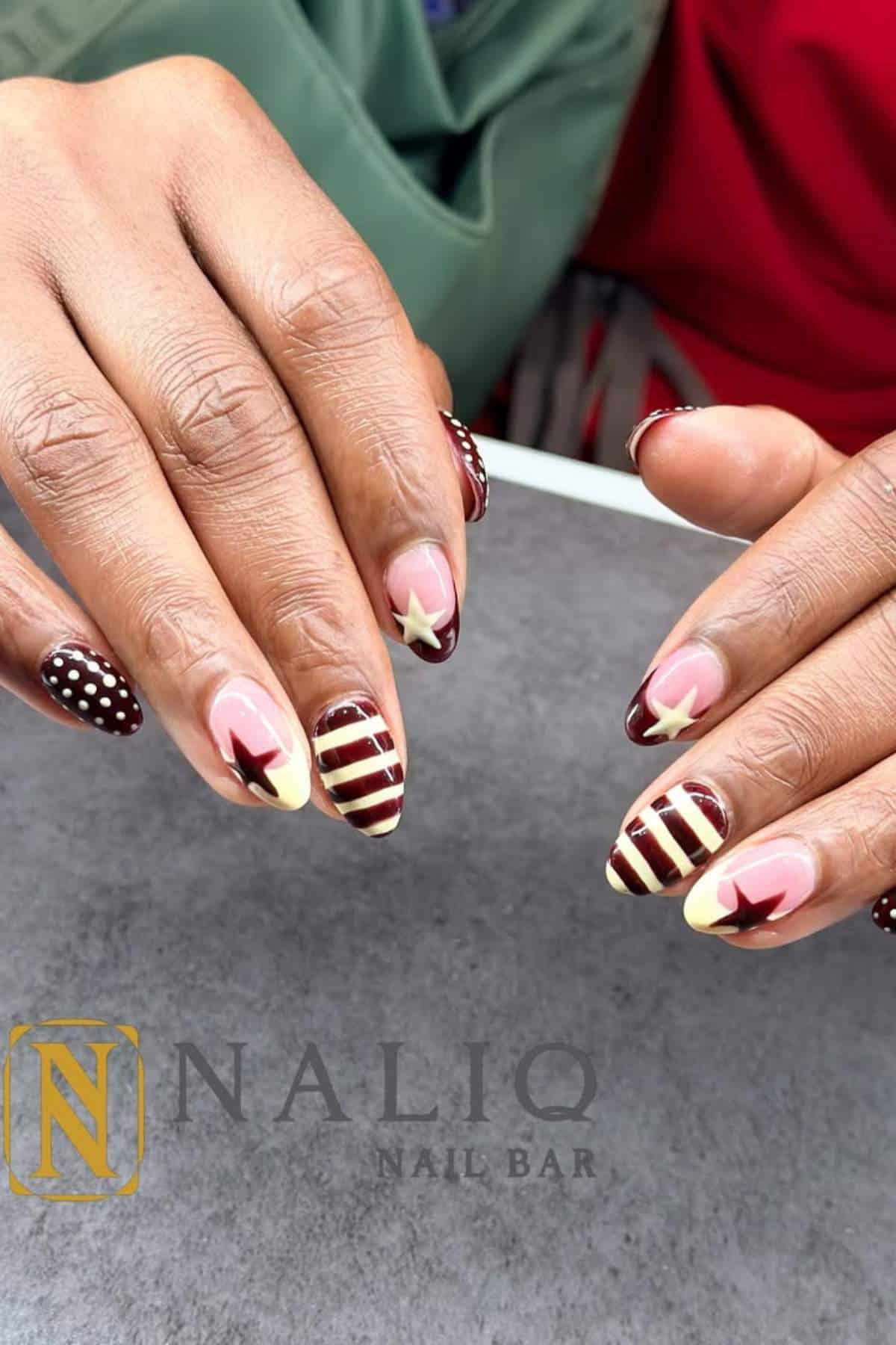 A person displays manicured nails with brown and cream stripes, stars, and polka dot designs, resting hands on a gray surface. NALIQ Nail Bar logo is visible.