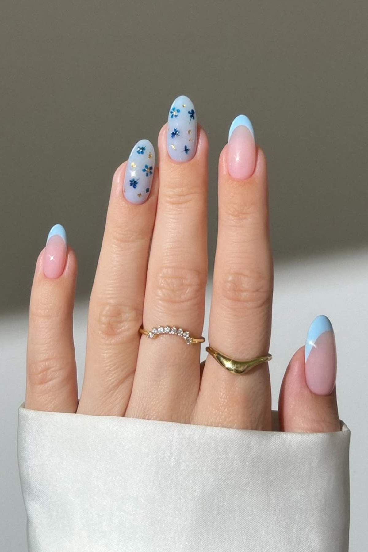 A hand with almond-shaped nails featuring a pastel blue French tip design. Some nails have blue floral patterns. The person is wearing two gold rings and a white sleeve.