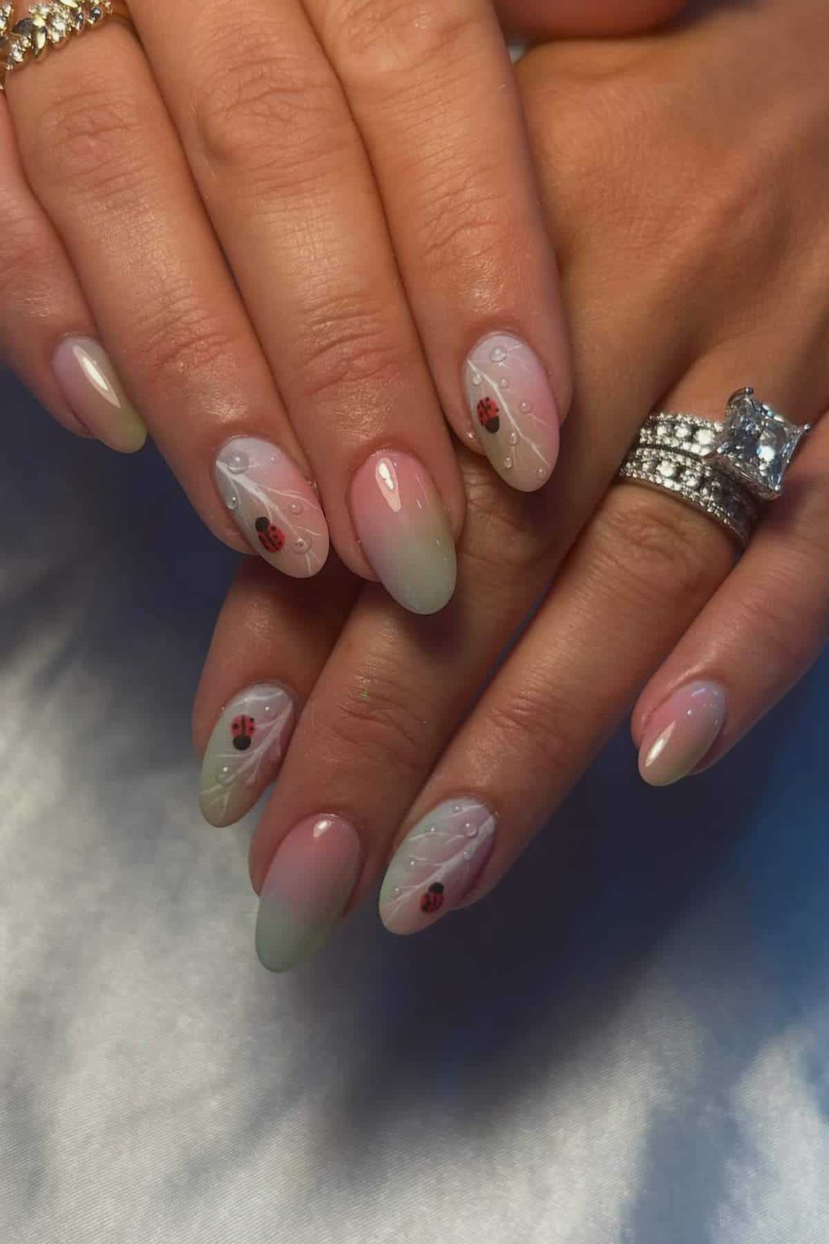 Hands with almond-shaped nails featuring a pale pink to green ombre, small ladybug designs, and white dot accents. The person is wearing two rings, including a large diamond ring.