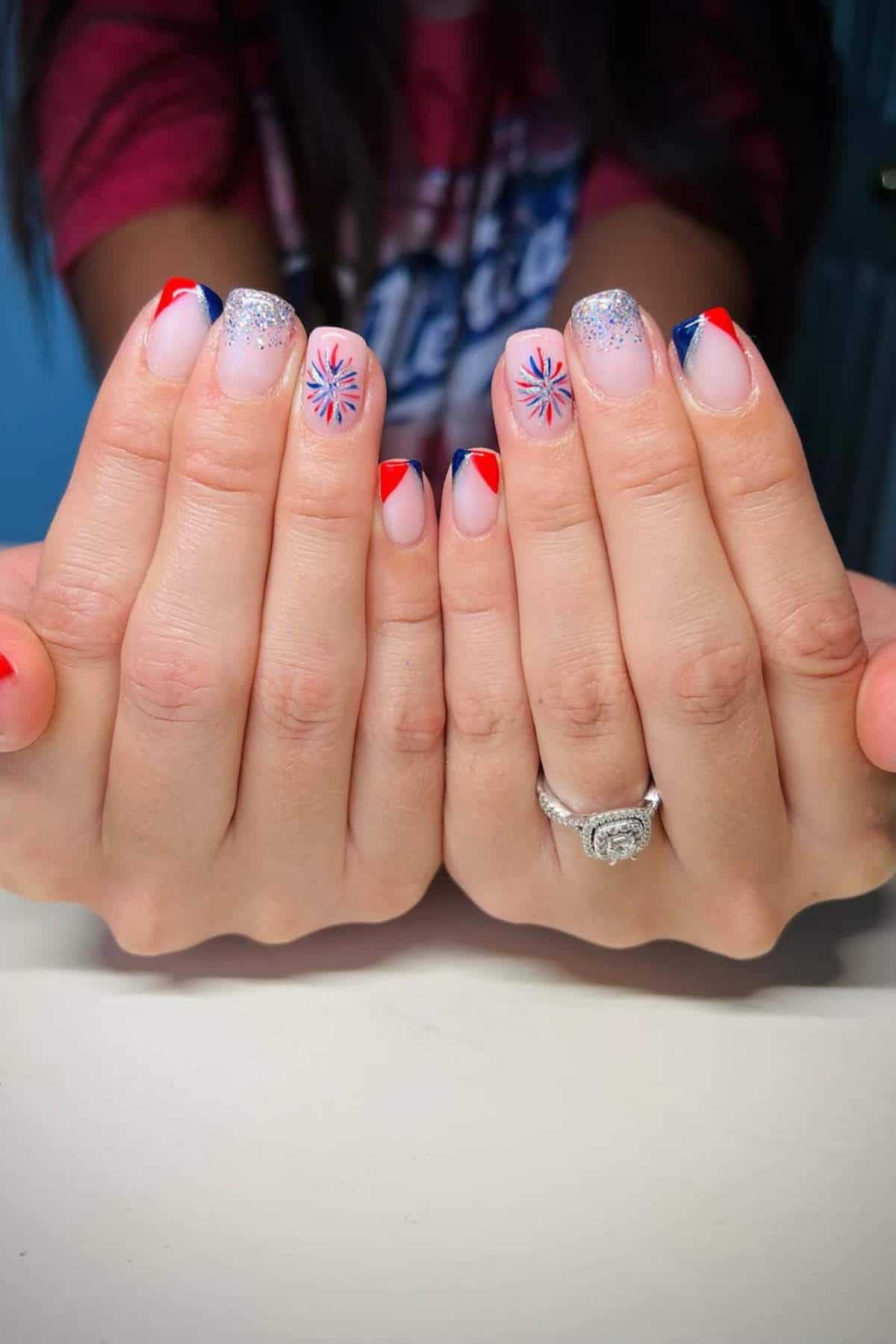 Close-up of hands with patriotic-themed nails featuring red, white, and blue designs, including fireworks and glitter accents.