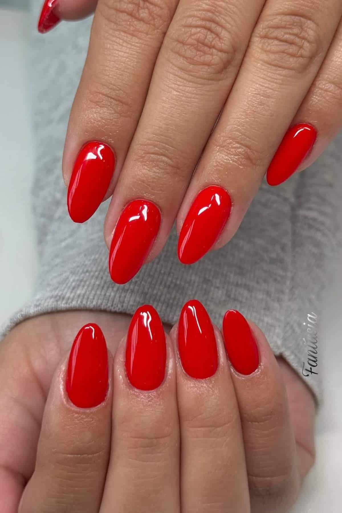 Close-up of hands with almond-shaped nails painted glossy bright red, resting on a light gray sleeve.