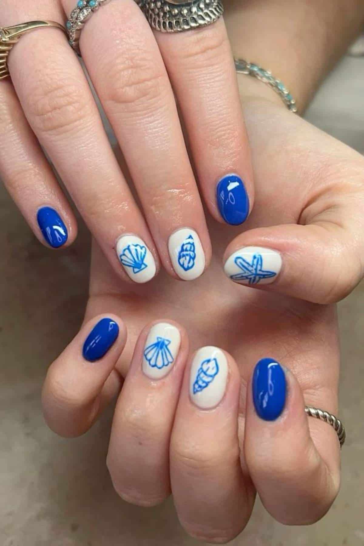 Hands with manicured nails painted in blue and white, featuring shell and starfish designs drawn in blue on the white nails.
