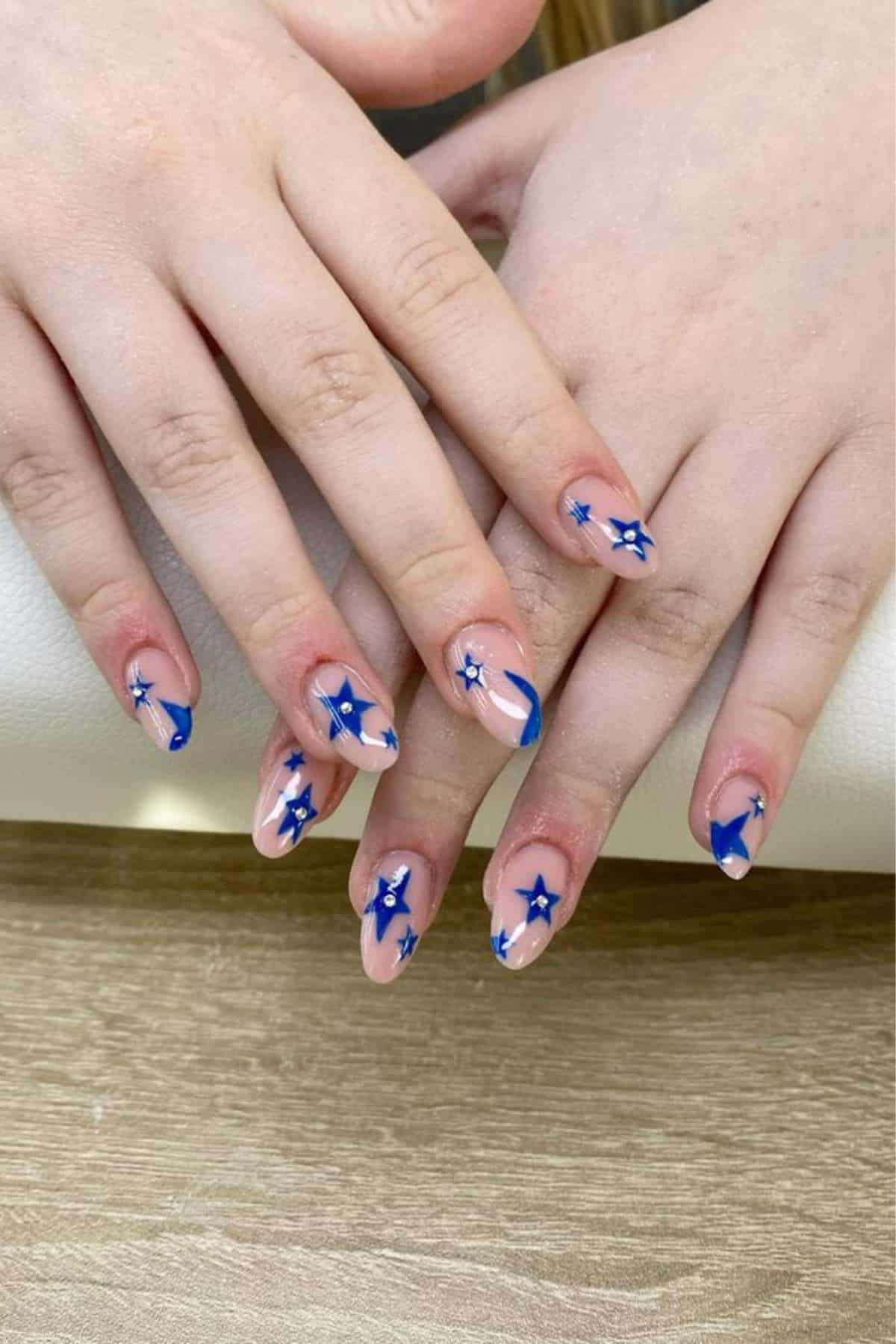 A pair of hands with almond-shaped nails featuring a nude base and blue star and swirl nail art designs.