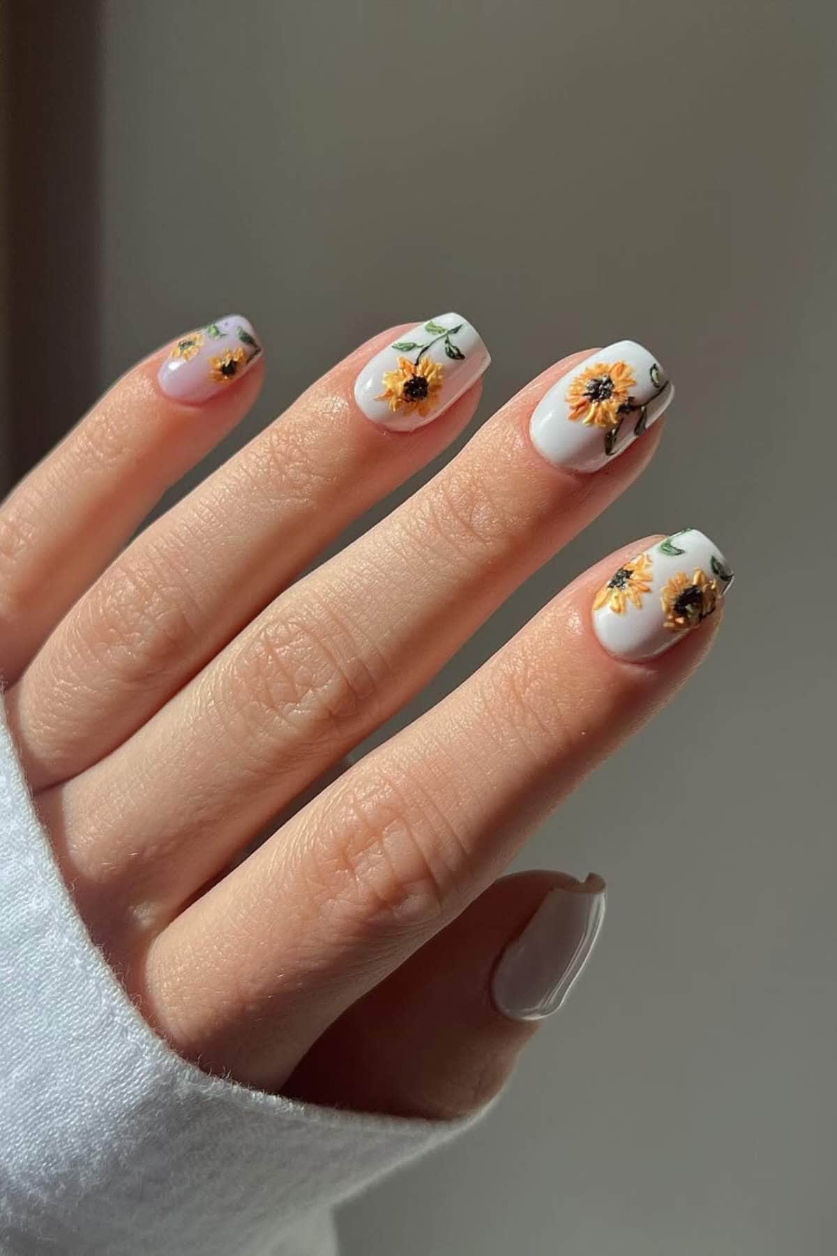 A hand with short, oval-shaped nails painted white and decorated with yellow and orange floral designs.