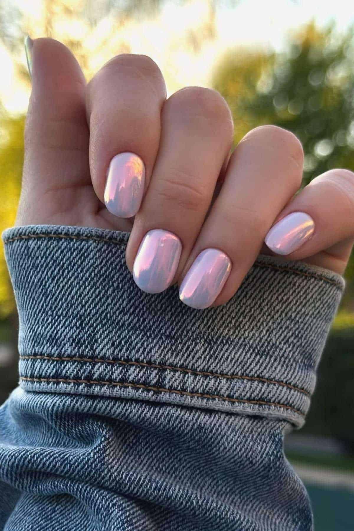 A hand with short, iridescent light pink nails is shown against a blurry outdoor background, wearing a blue denim jacket.