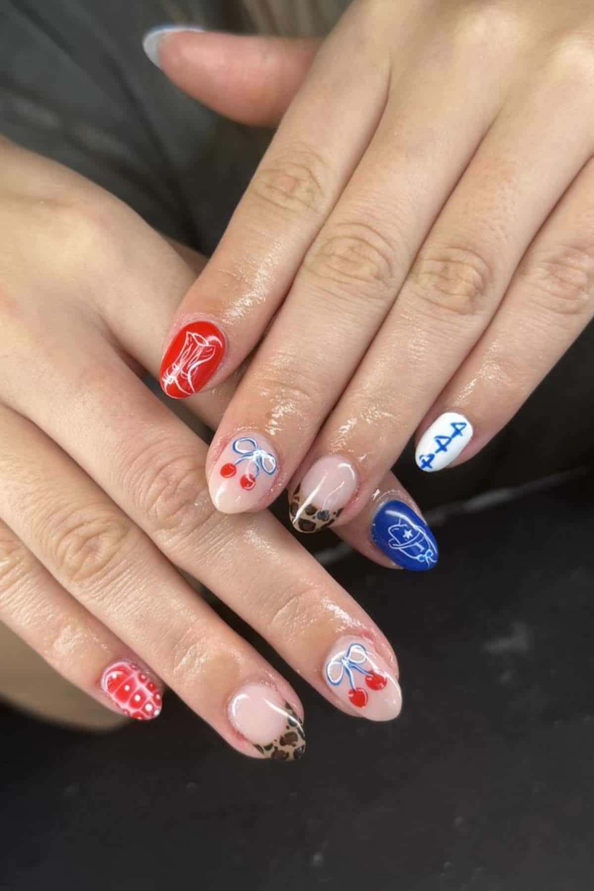 Hands with manicured nails featuring various designs, including cherries, a red sneaker, blue hearts, red polka dots, leopard print, and a blue stitched heart.