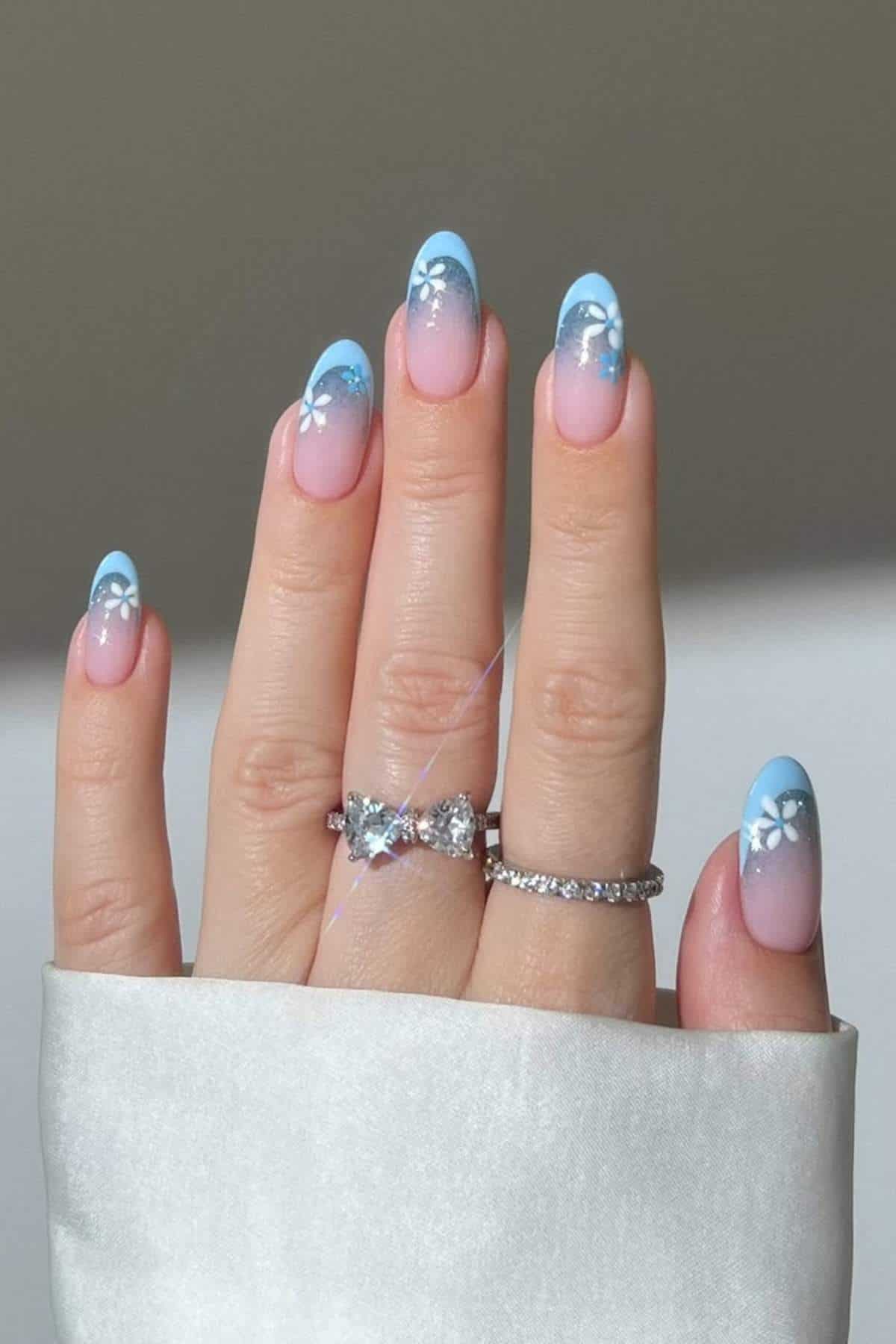 A hand with long, almond-shaped nails painted in a blue to pink ombre gradient with white floral designs, wearing two silver rings on the ring finger.