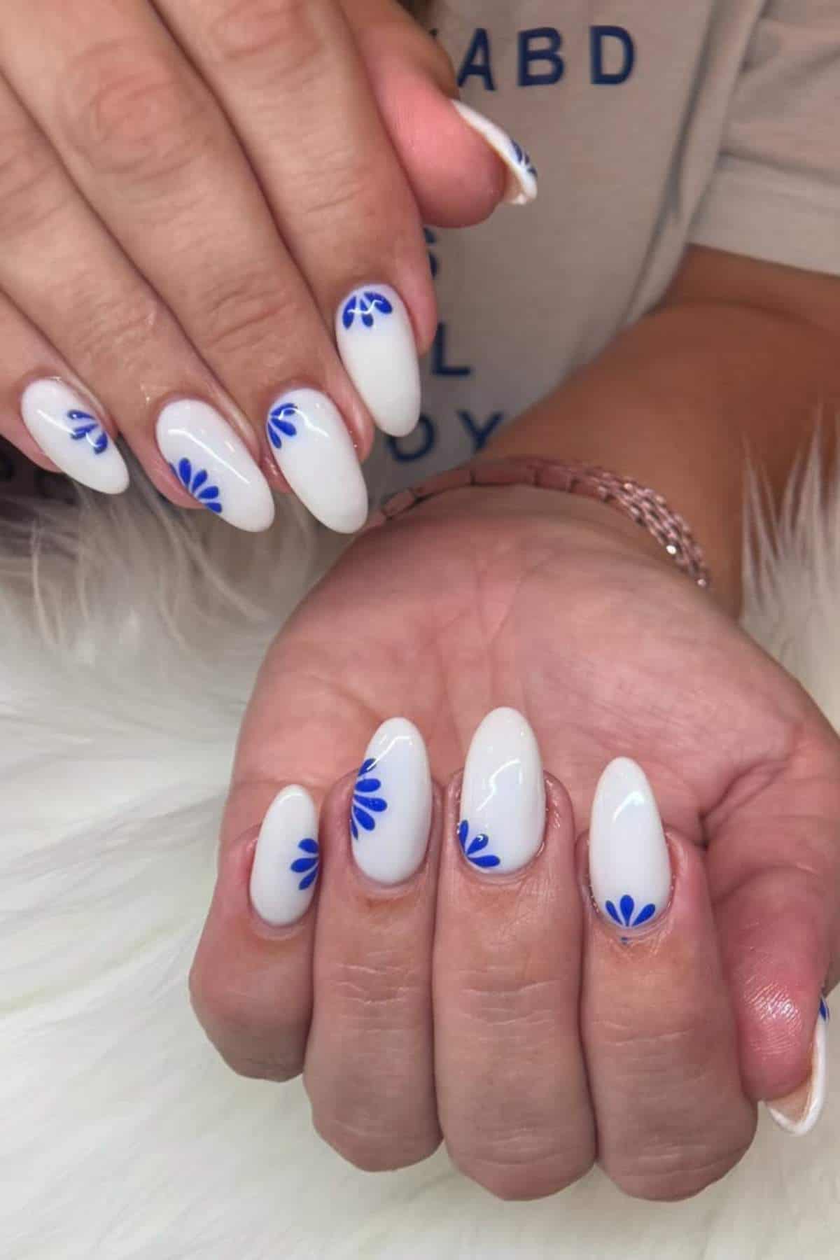 Hands with almond-shaped white nails featuring small, blue abstract flower designs near the cuticles, displayed on a white surface.