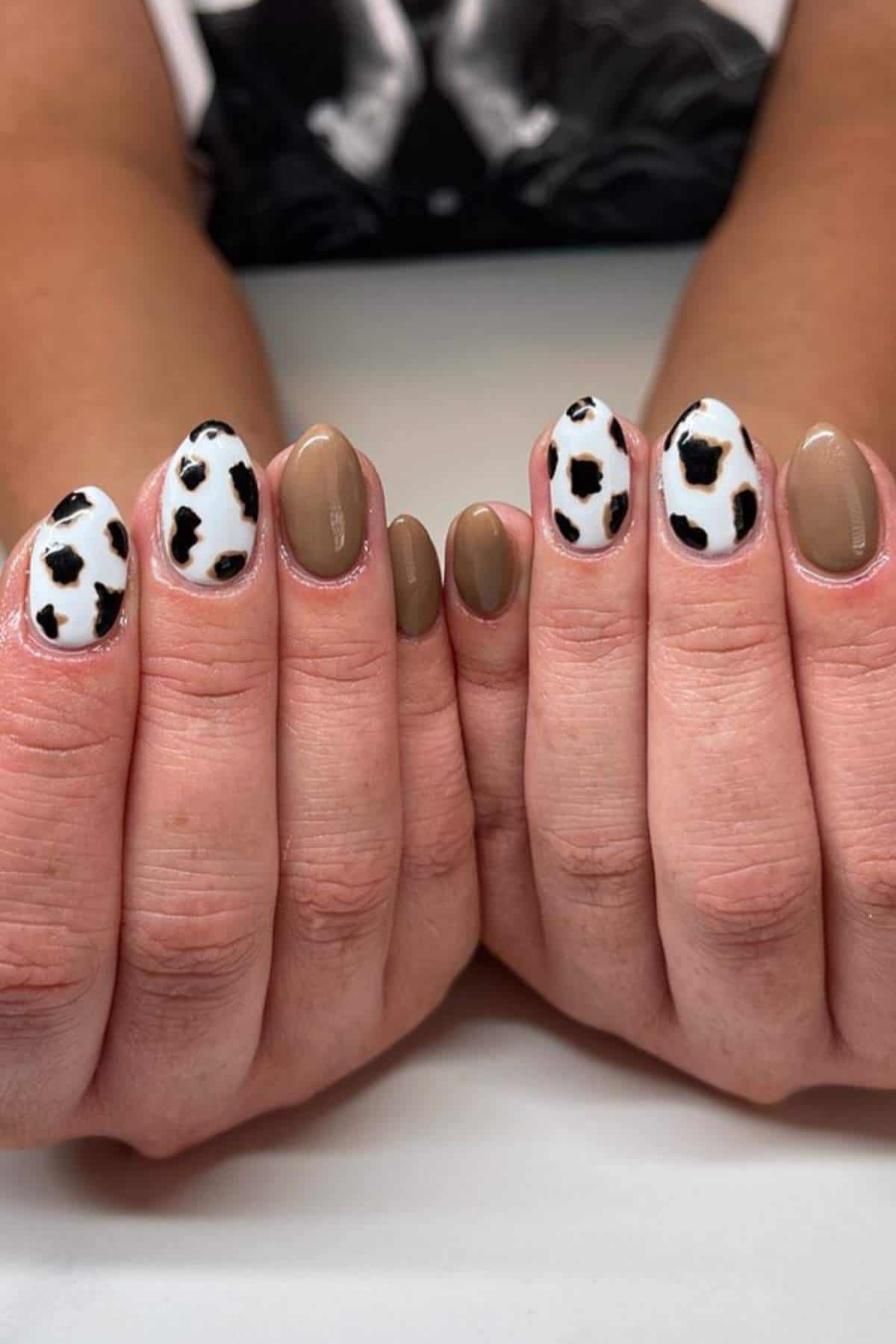 Two hands with short almond-shaped nails; some nails are painted solid beige while others have a white base with black cow print spots.