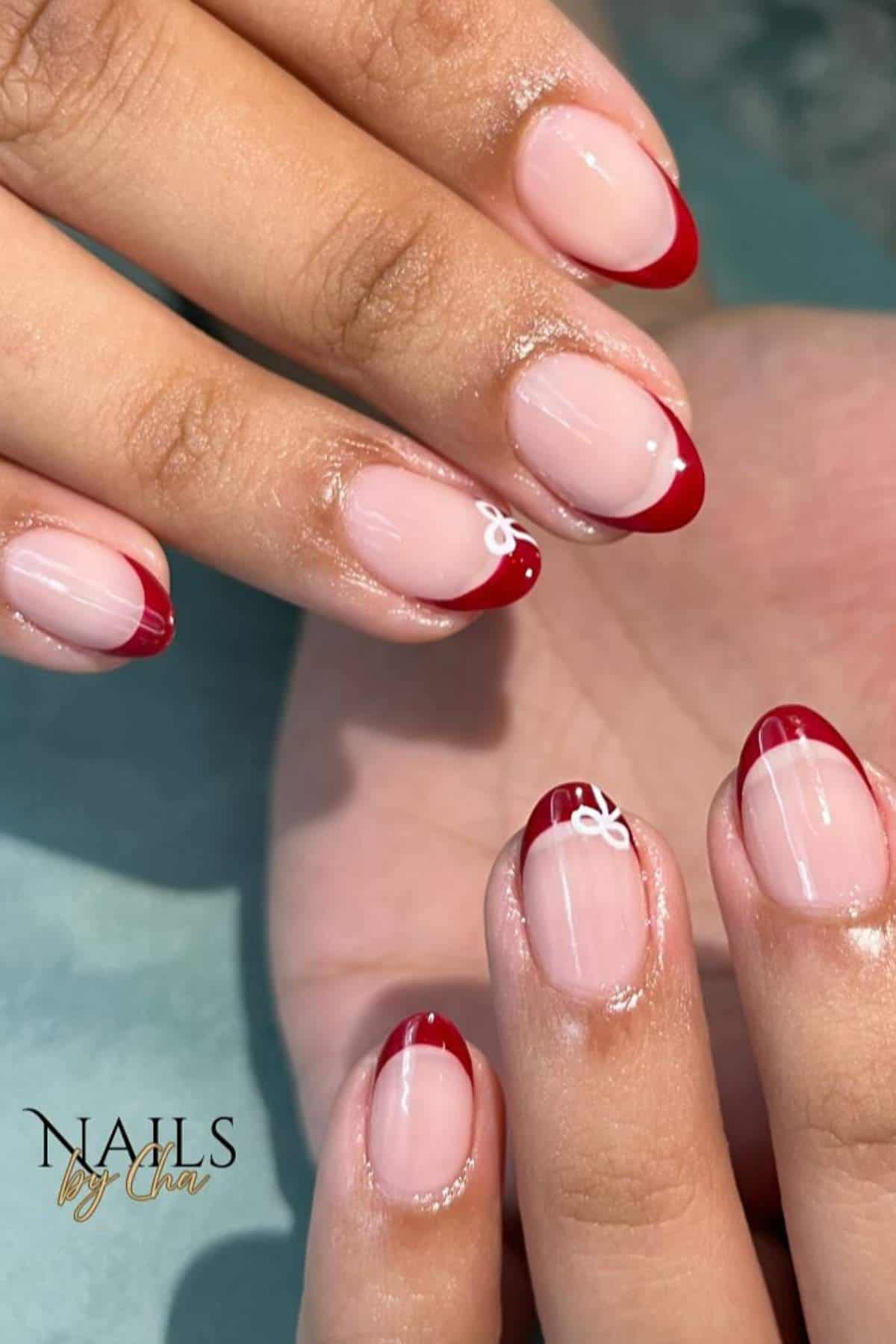 Hands with almond-shaped nails featuring a red French tip manicure; some nails have a small white bow design near the tips. "Nails by Cha" logo in the lower left corner.