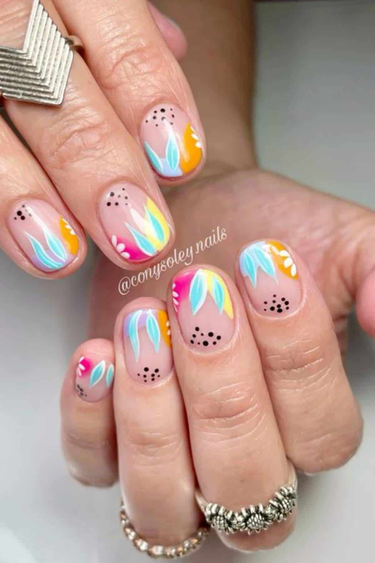 Close-up of hands with nude nail polish featuring colorful abstract leaf designs in blue, yellow, pink, and orange, plus dotted black accents.