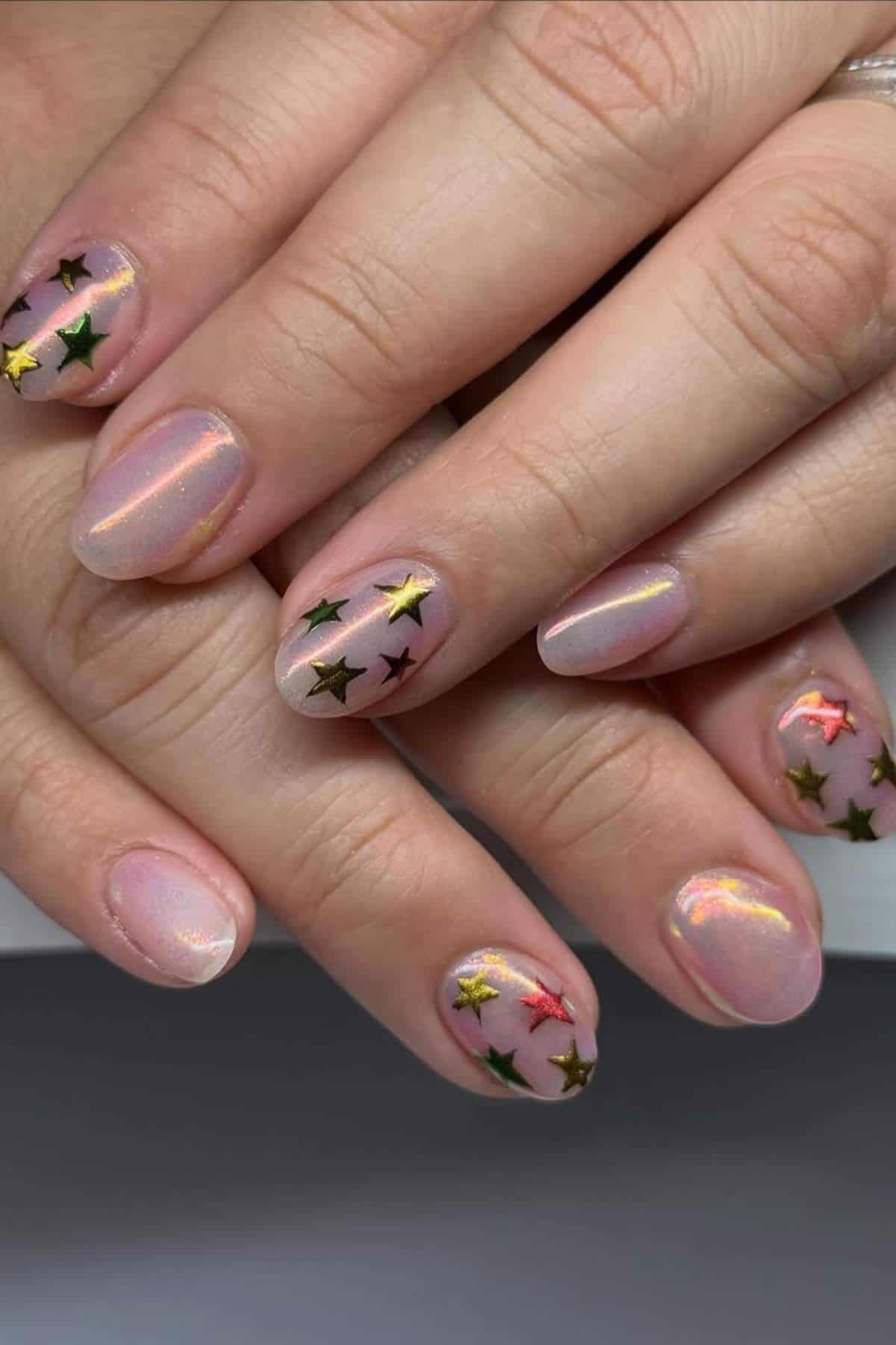 Close-up of hands with short, round nails painted in a sheer nude polish, decorated with gold and green metallic star-shaped nail art stickers.