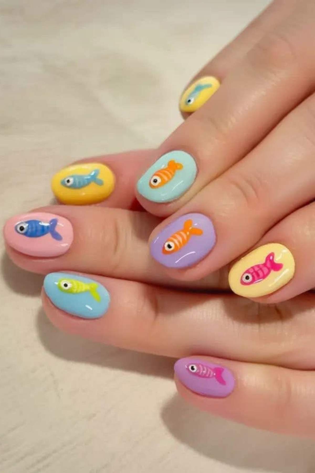 Close-up of hands with pastel-colored nail polish and small, colorful fish designs on each fingernail.