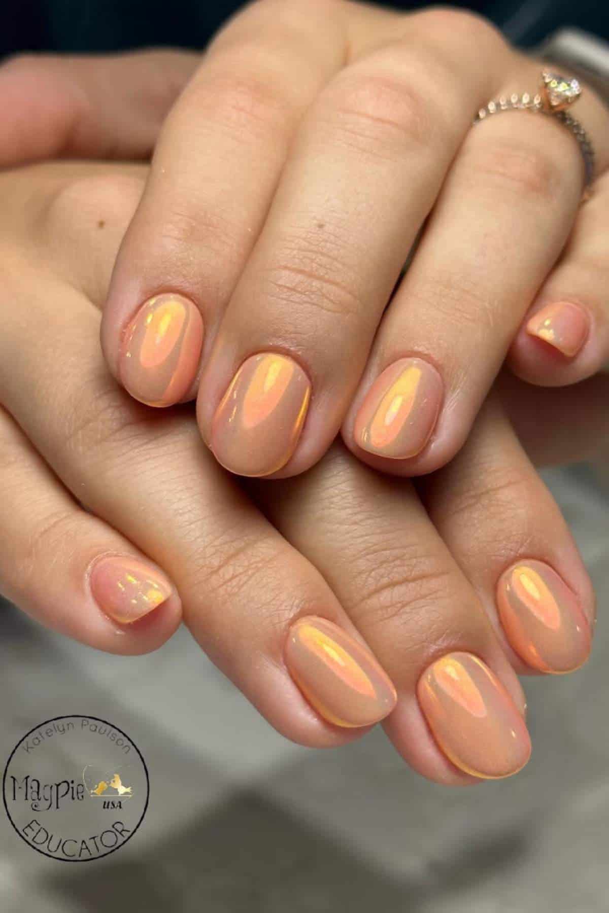Close-up of hands with short, almond-shaped nails painted in a glossy peach-orange polish with a subtle iridescent shimmer.