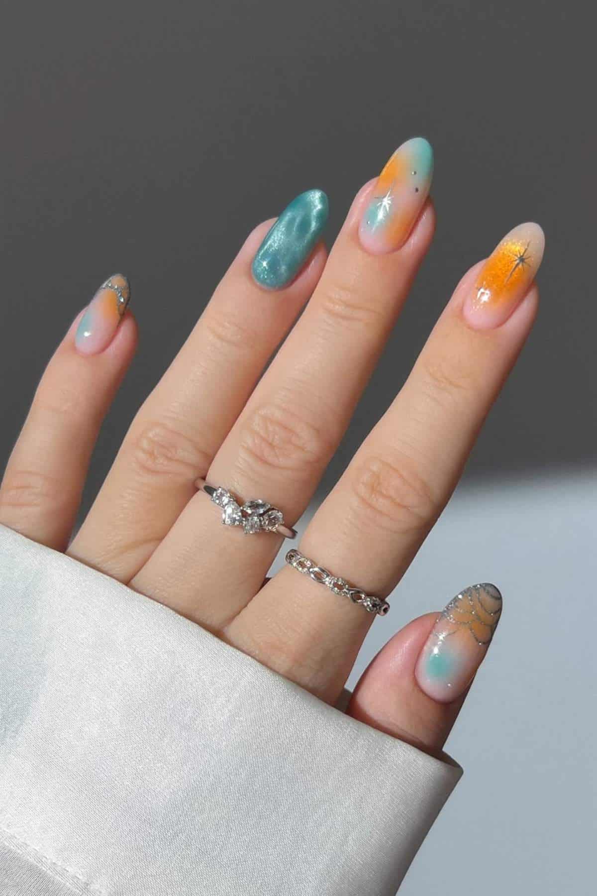 A hand with long, almond-shaped nails featuring blue, orange, and yellow gradient nail art with silver star and glitter details; wearing three silver rings.