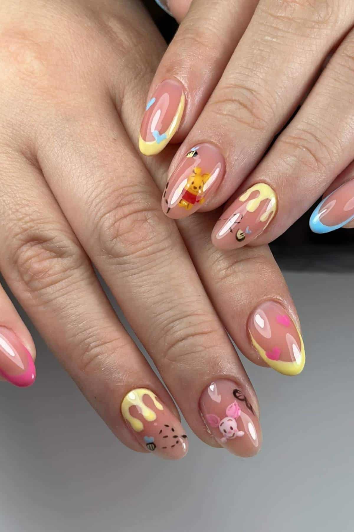 Close-up of hands with almond-shaped nails featuring pastel yellow, pink, and blue tips, honey drip designs, and cartoon characters resembling Winnie the Pooh and Piglet.