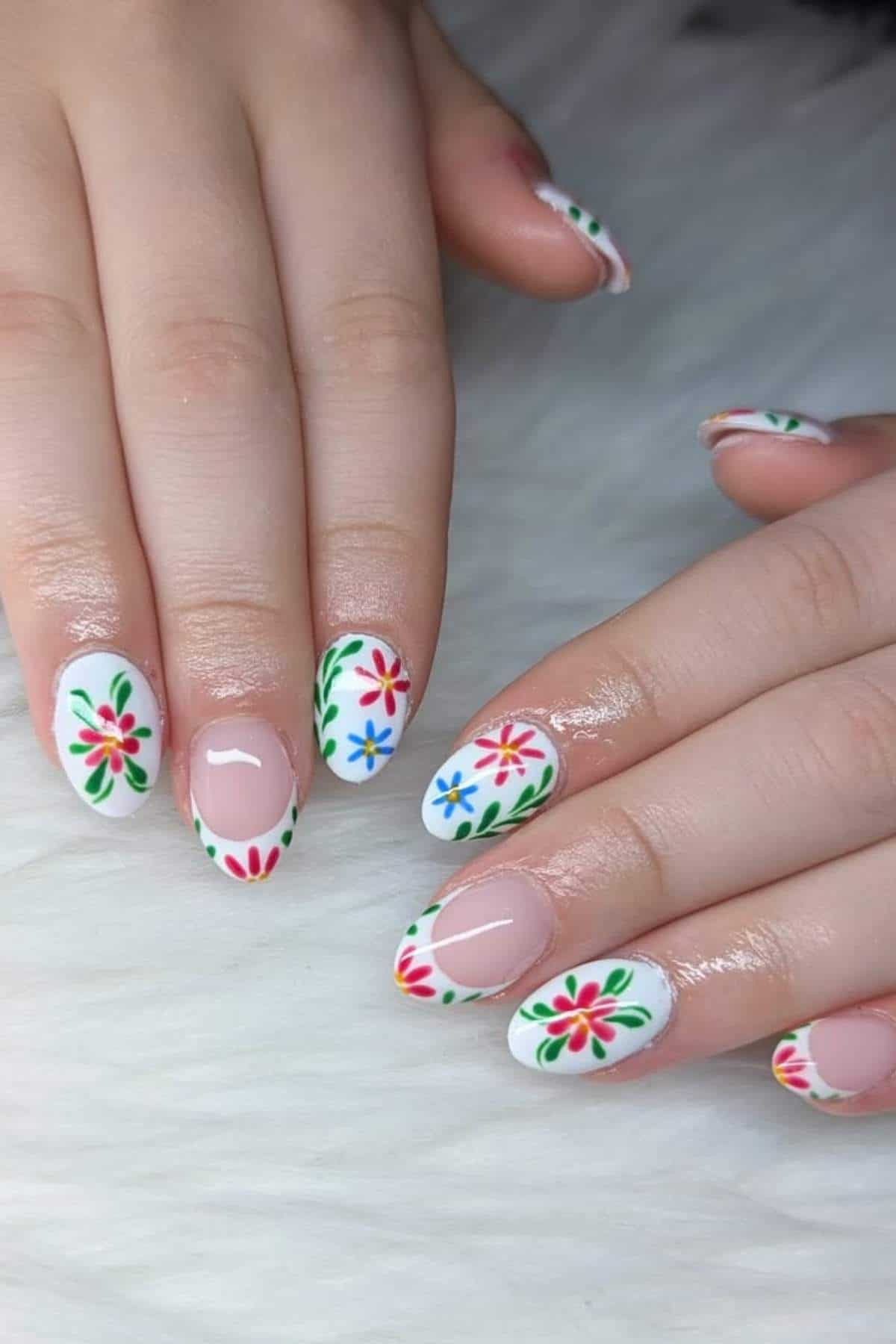Hands with almond-shaped nails featuring white tips and colorful floral designs in red, green, and blue, resting on a white textured surface.