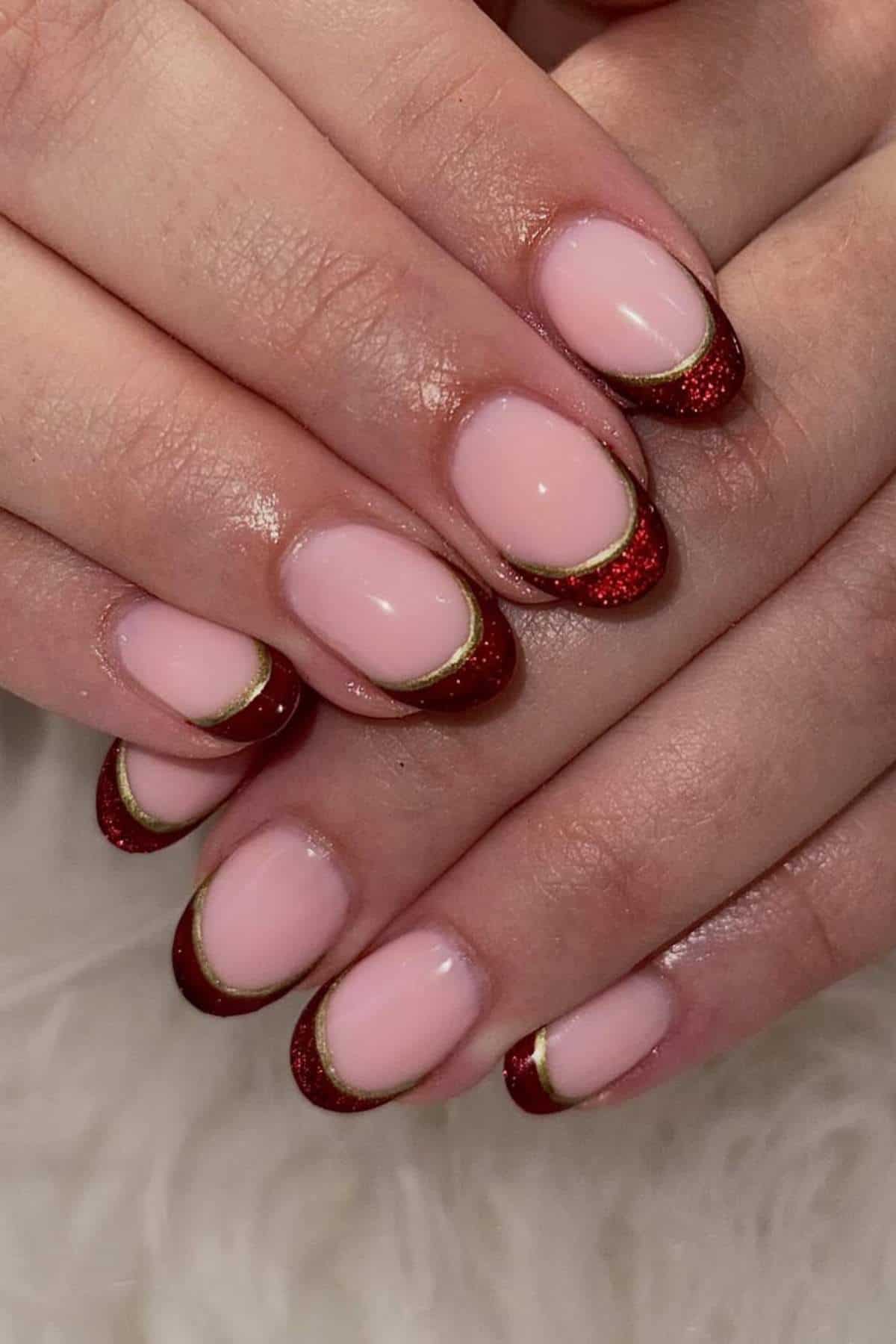 Hands with manicured nails featuring a pale pink base, red glitter tips, and thin gold line accents along the French tip edges.