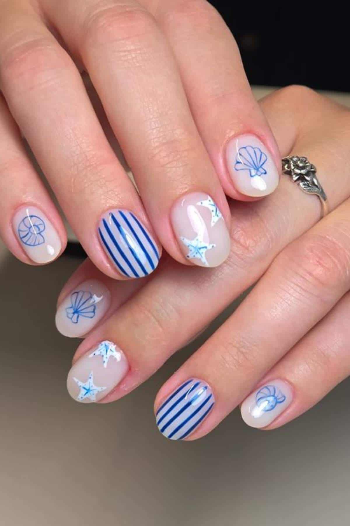 A hand with neatly manicured nails featuring blue seashells, white starfish, and vertical blue stripes on a nude base.