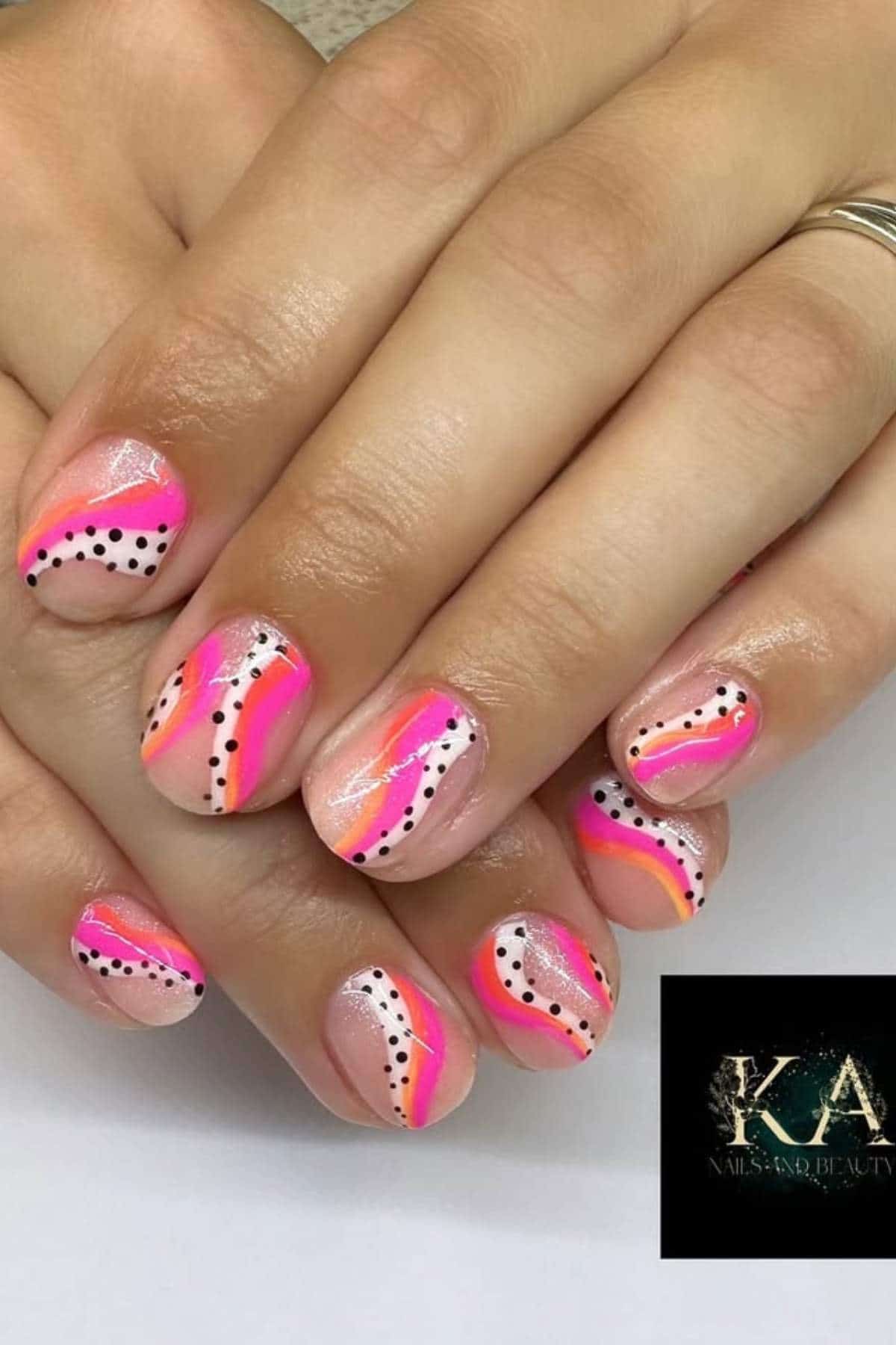 Hands with short, rounded nails featuring a colorful abstract design in pink, orange, and white, accented with black dotted lines. A "KA Nails and Beauty" logo is visible in the corner.