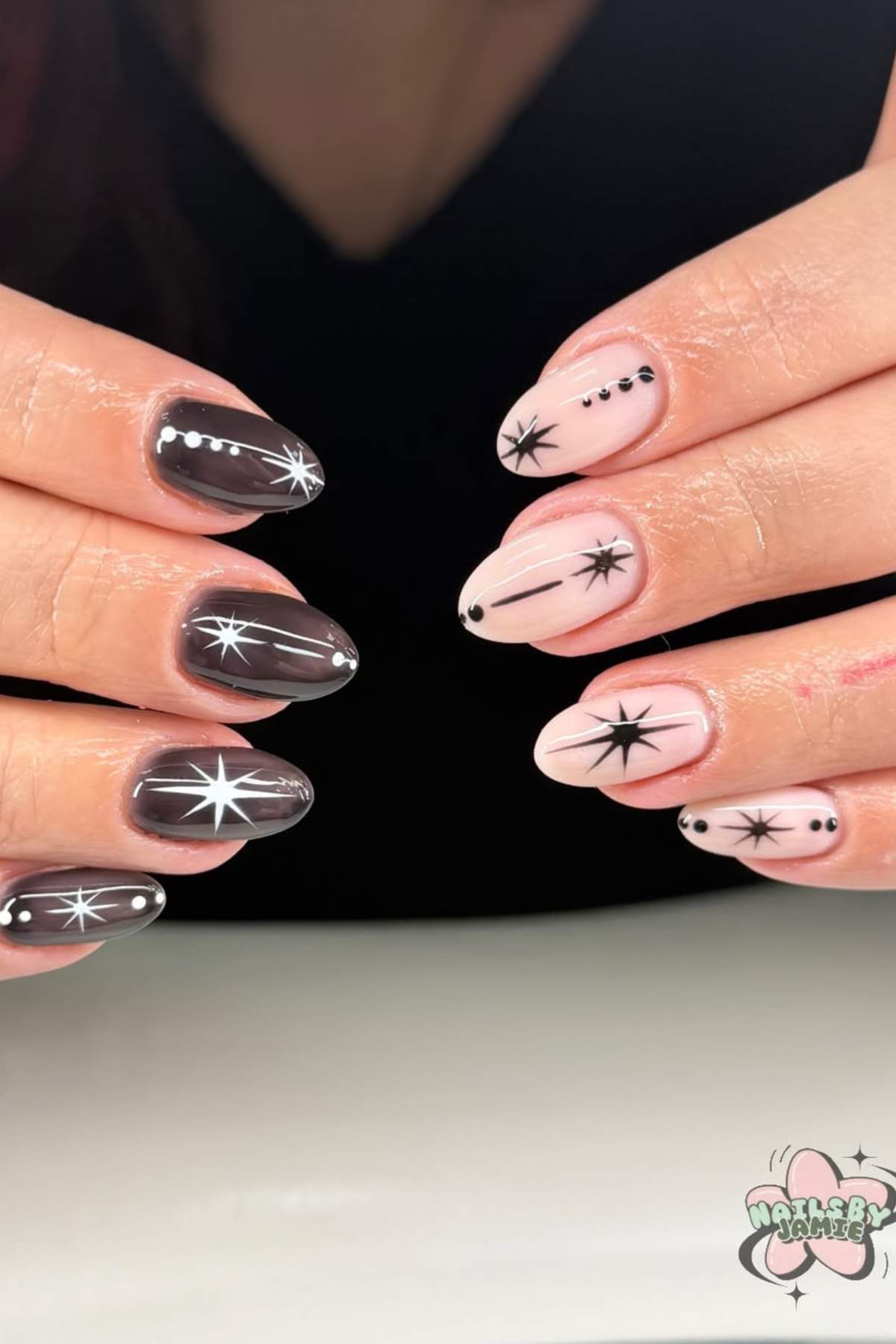 Two hands with almond-shaped nails, painted in black and pale pink shades, featuring white celestial star and dot designs on each nail.