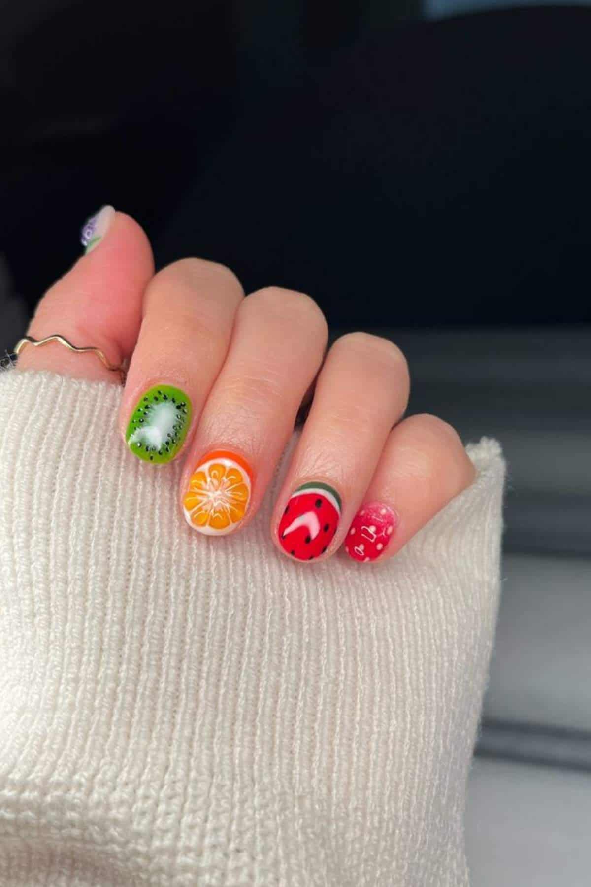 A hand with fruit-themed nail art, including designs of kiwi, orange, watermelon, and strawberry, partially covered by a cream-colored sleeve.