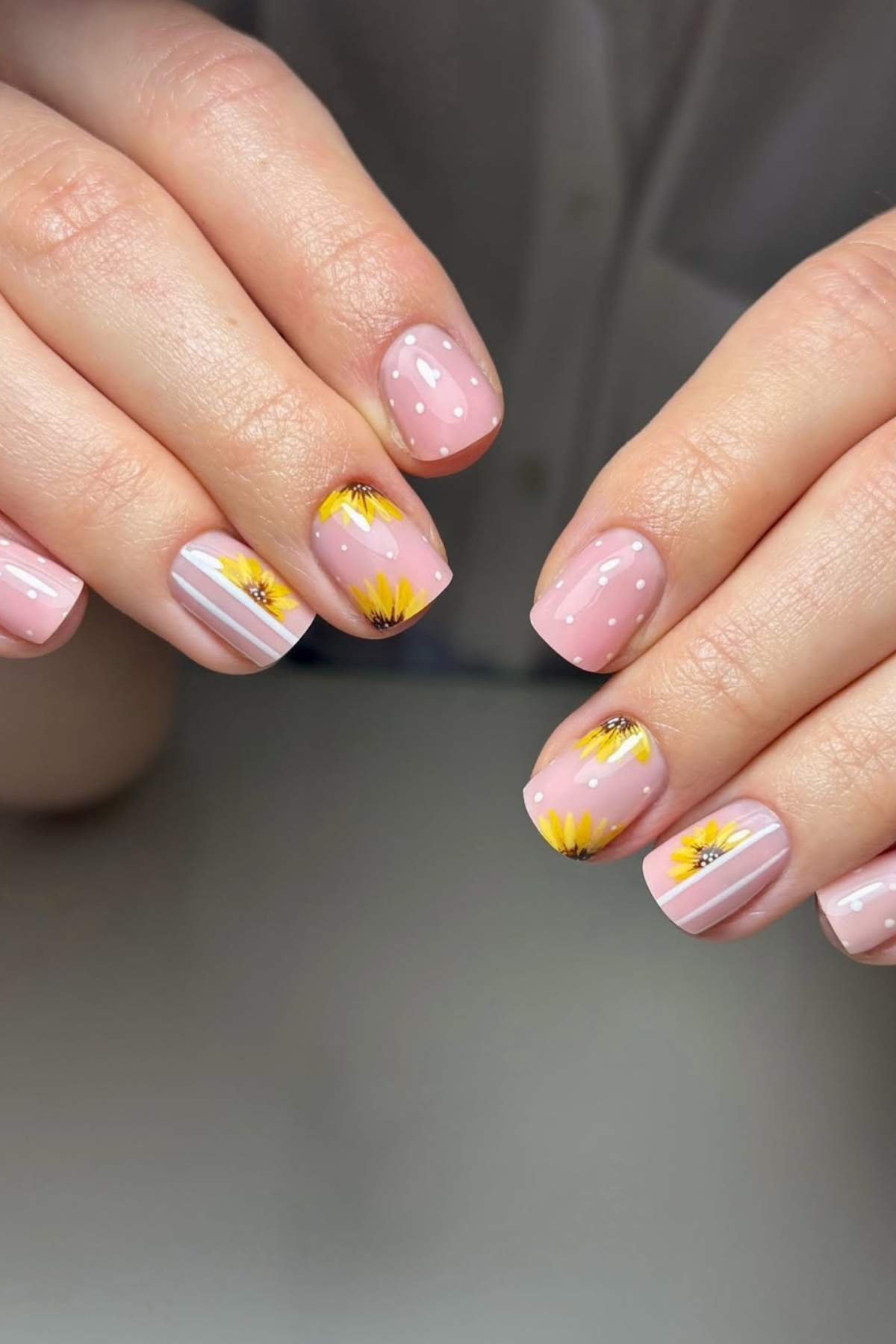 Close-up of hands with short, light pink nails featuring painted yellow sunflowers and white accent lines on a few nails.