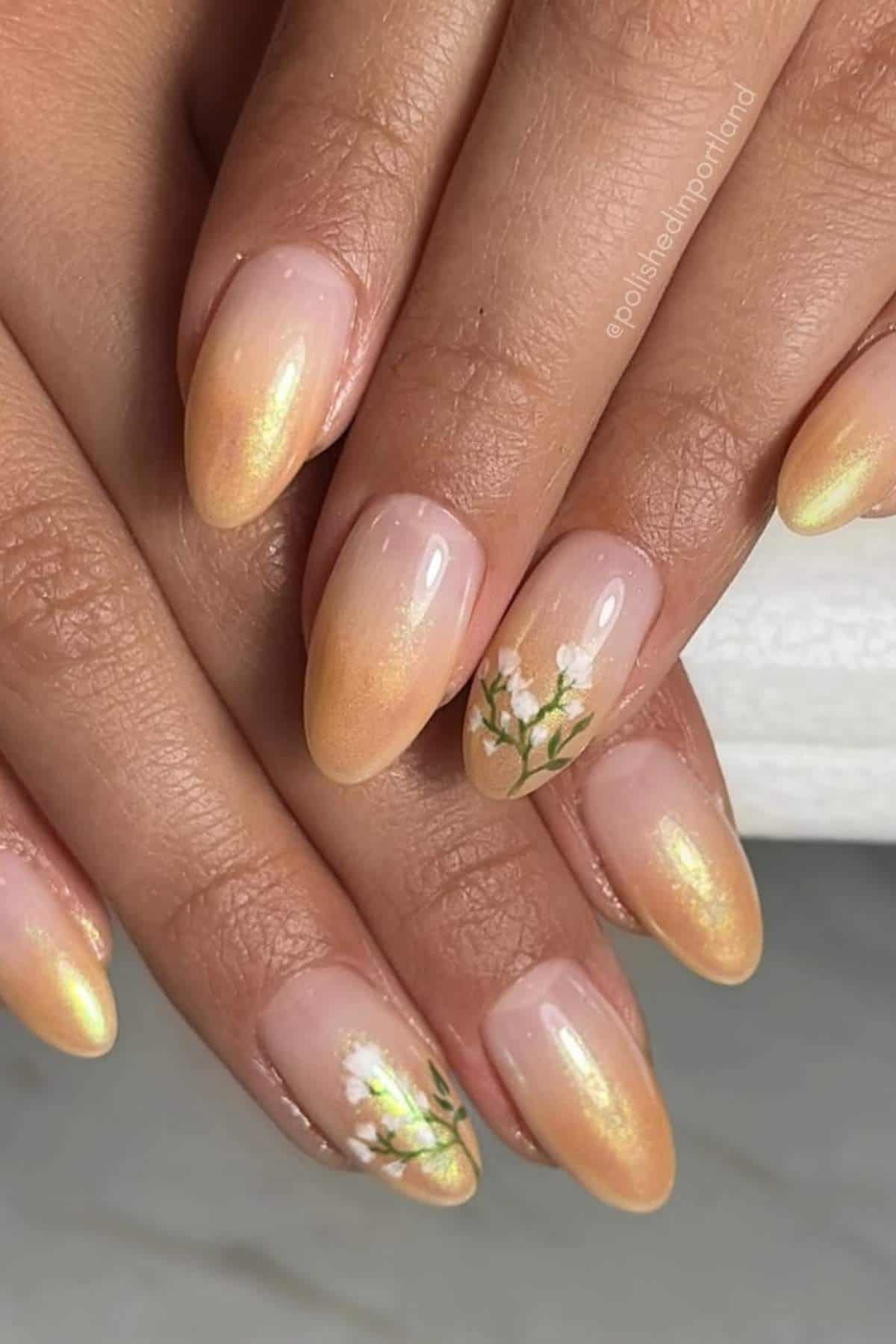 Hands with almond-shaped nails painted in a gold ombre style, featuring small white floral designs on the ring fingers.