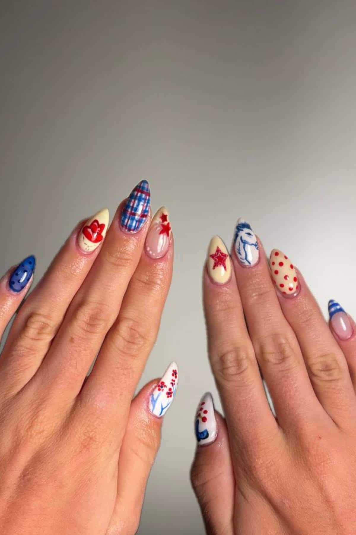 Two hands with almond-shaped nails featuring various red, white, and blue designs, including stars, plaid, dots, hearts, and abstract patterns on each nail.