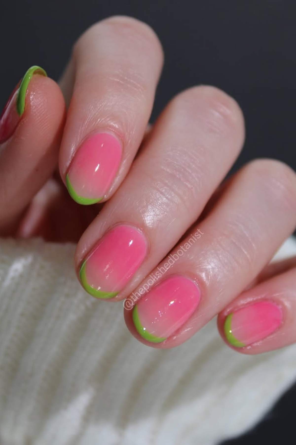 A hand with pink nails featuring green tips, shown against a dark background and partially covered by a white sweater sleeve.