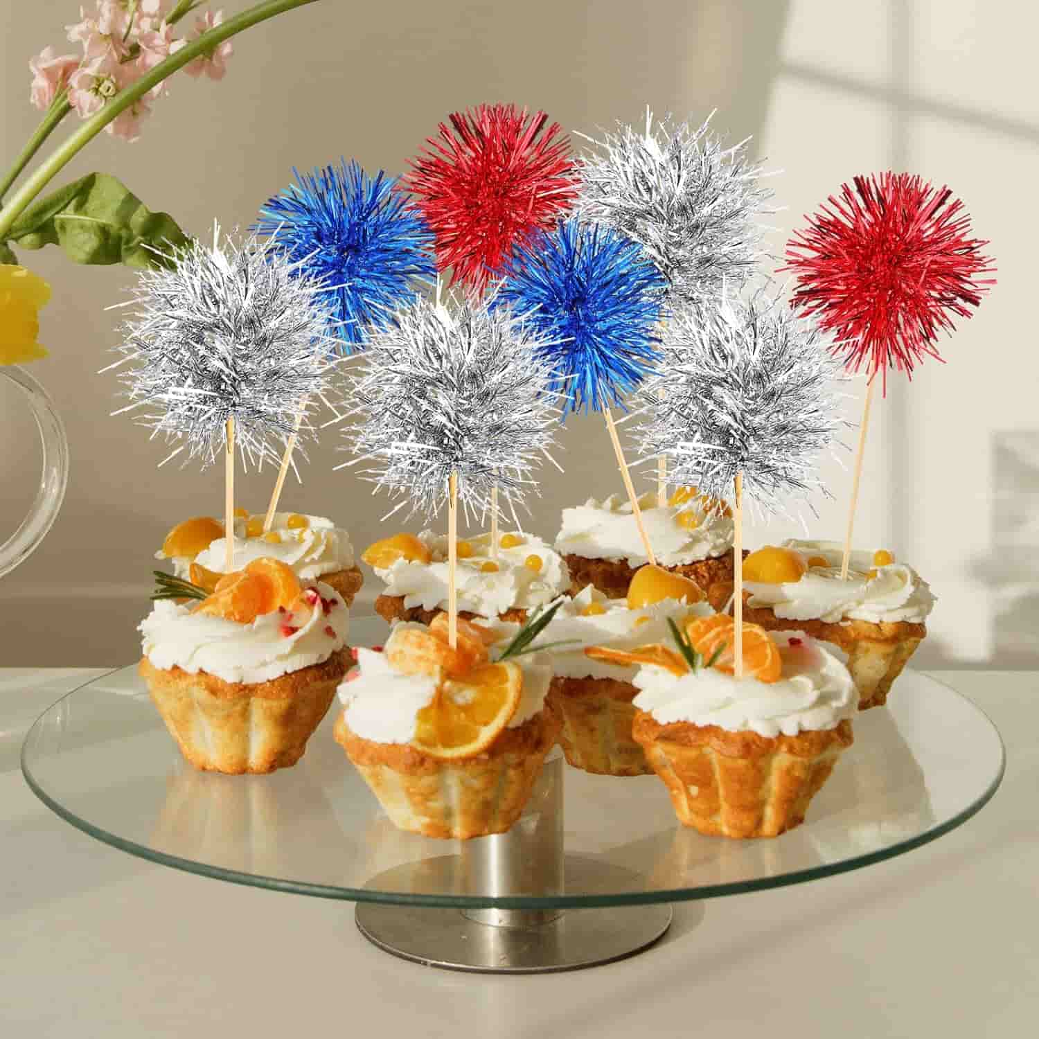 Cupcakes on a glass stand, each topped with whipped cream, orange slices, and red, blue, or silver tinsel pom-pom decorations.