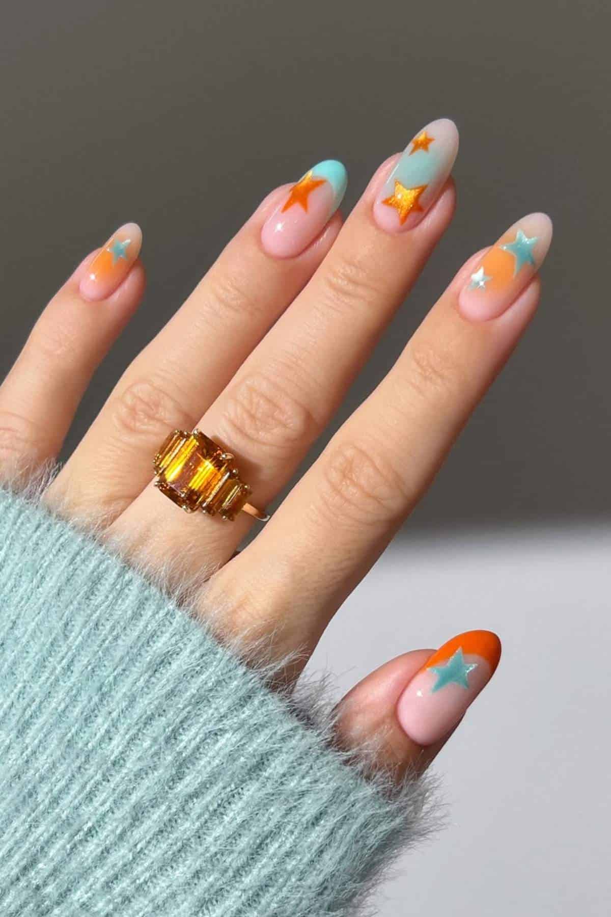 A hand with almond-shaped nails painted in pastel colors and decorated with colorful star designs, wearing a gold ring and a light blue fuzzy sweater.