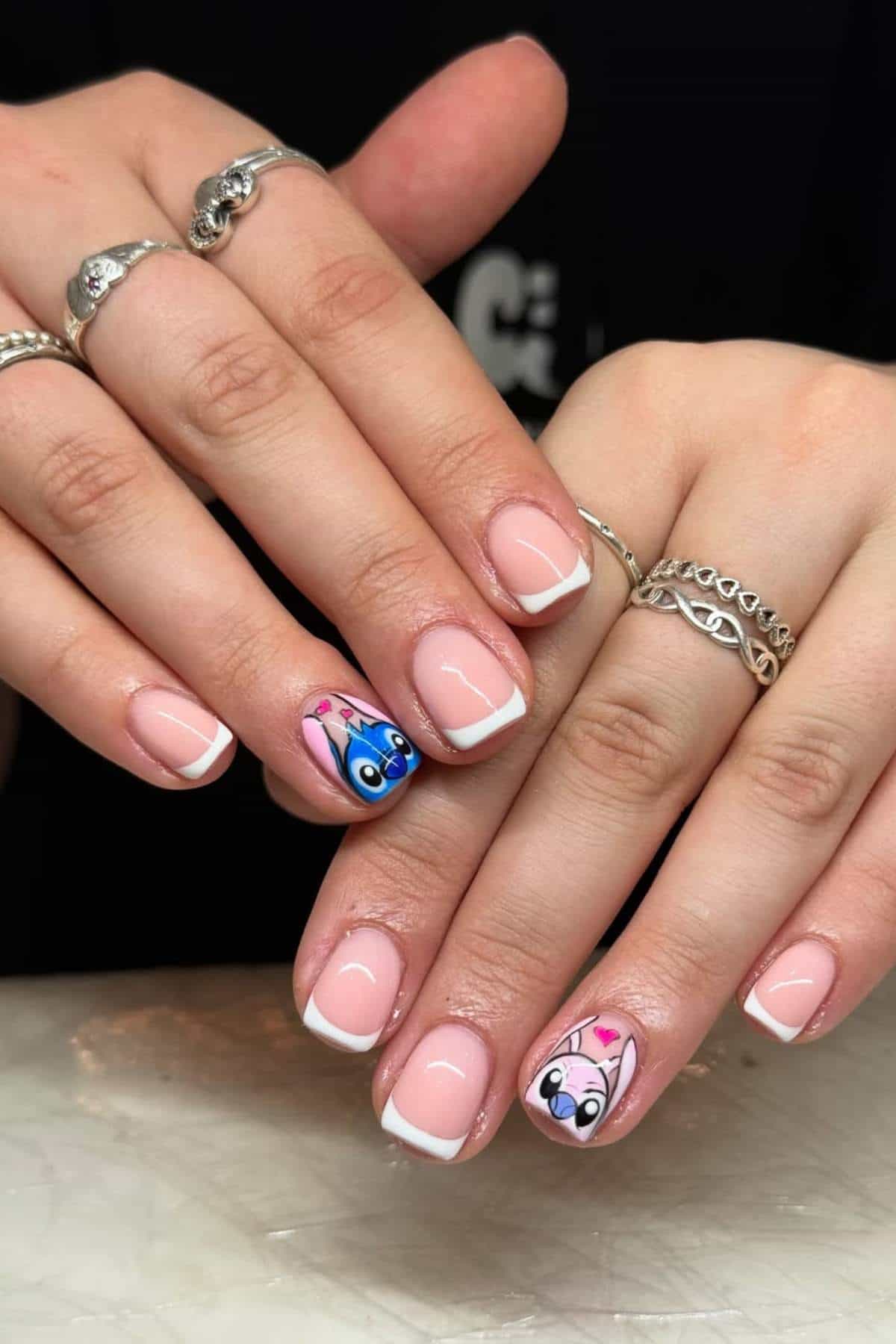 Hands with a French manicure; ring fingers feature cartoon character nail art with blue and white faces and pink hearts.