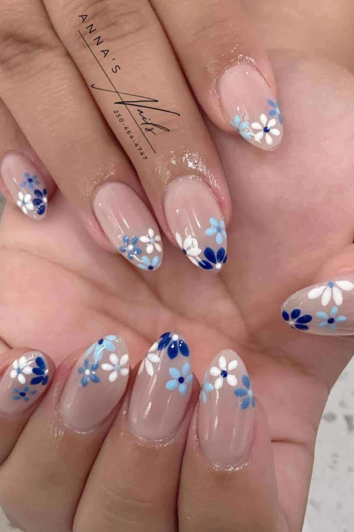 Close-up of hands with almond-shaped nails featuring a clear base and blue and white daisy flower nail art on the tips.