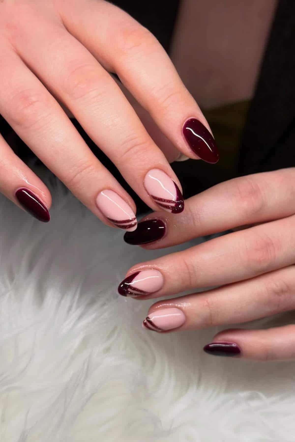 Close-up of hands with almond-shaped nails featuring a mix of solid burgundy polish and pink nails with burgundy tips and wavy accents, displayed on a white textured surface.