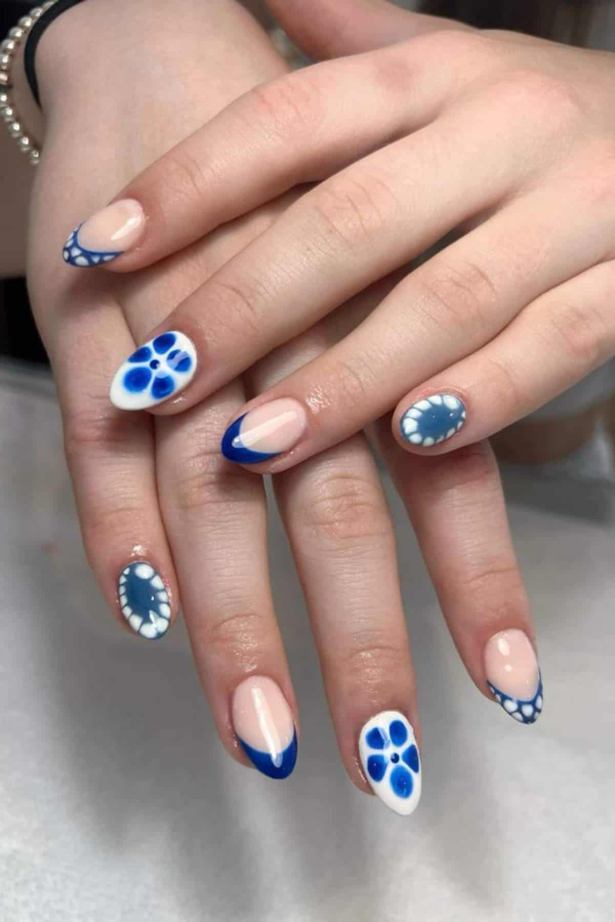 Hands with almond-shaped nails featuring blue and white designs, including floral patterns, polka dots, and French tips on a neutral base.