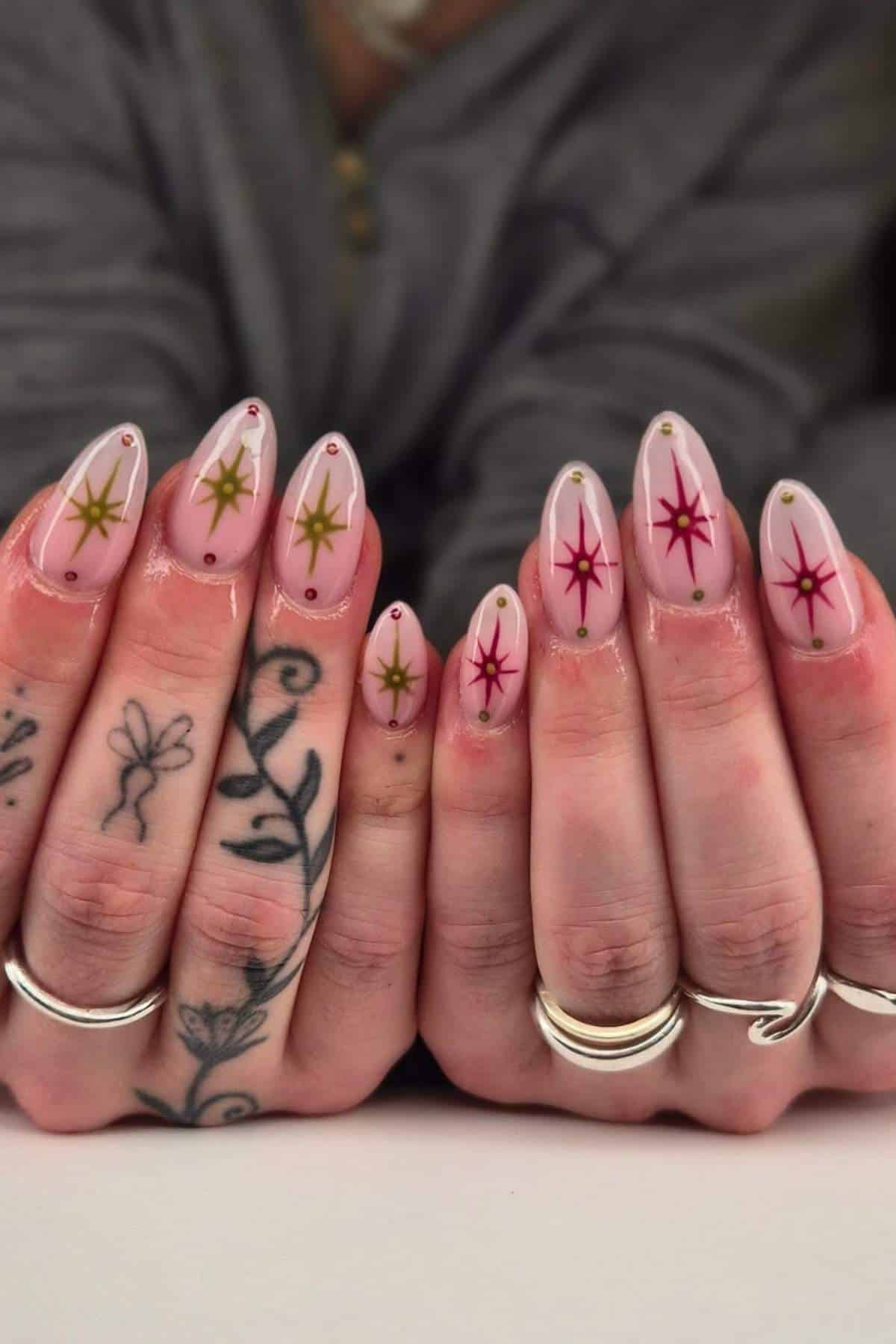 Hands with almond-shaped nails painted light pink, decorated with gold and magenta starburst designs and small gold studs; fingers adorned with silver rings and floral tattoos.