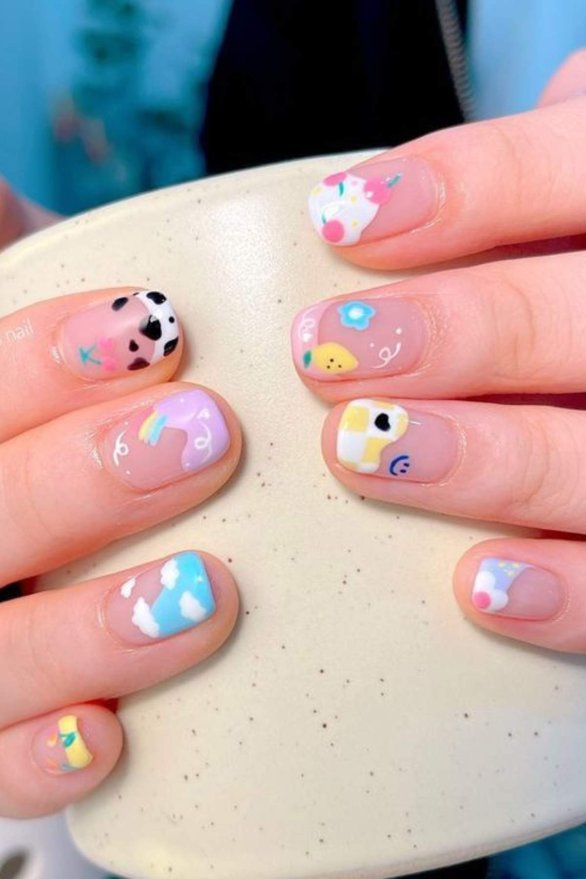 Close-up of hands with short nails painted in colorful, whimsical designs including clouds, rainbows, cow print, fruit, and abstract shapes on a neutral background.