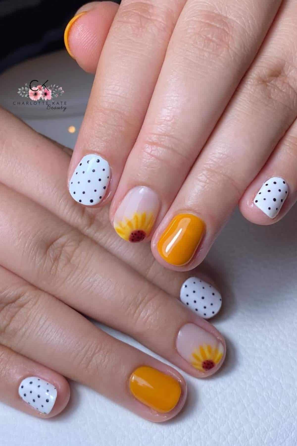 Close-up of hands with manicured nails featuring white with black polka dots, solid yellow, and yellow sunflower designs on a light background.