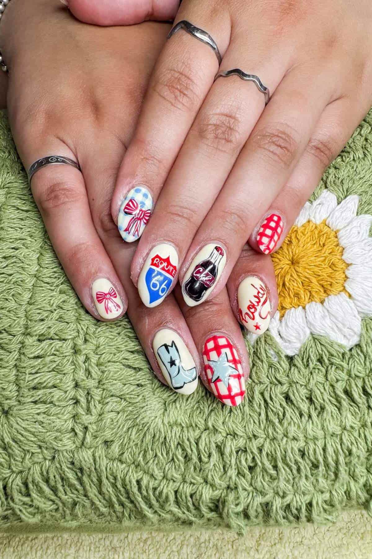 A hand with detailed nail art designs, including Route 66, bows, a Coke bottle, and checkered patterns, rests on a green fabric with a white and yellow flower.