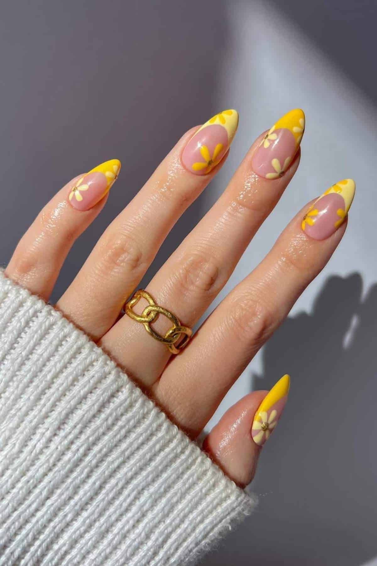 A hand with almond-shaped nails featuring yellow and white floral designs, wearing a chunky gold ring, and a white ribbed sweater sleeve visible.