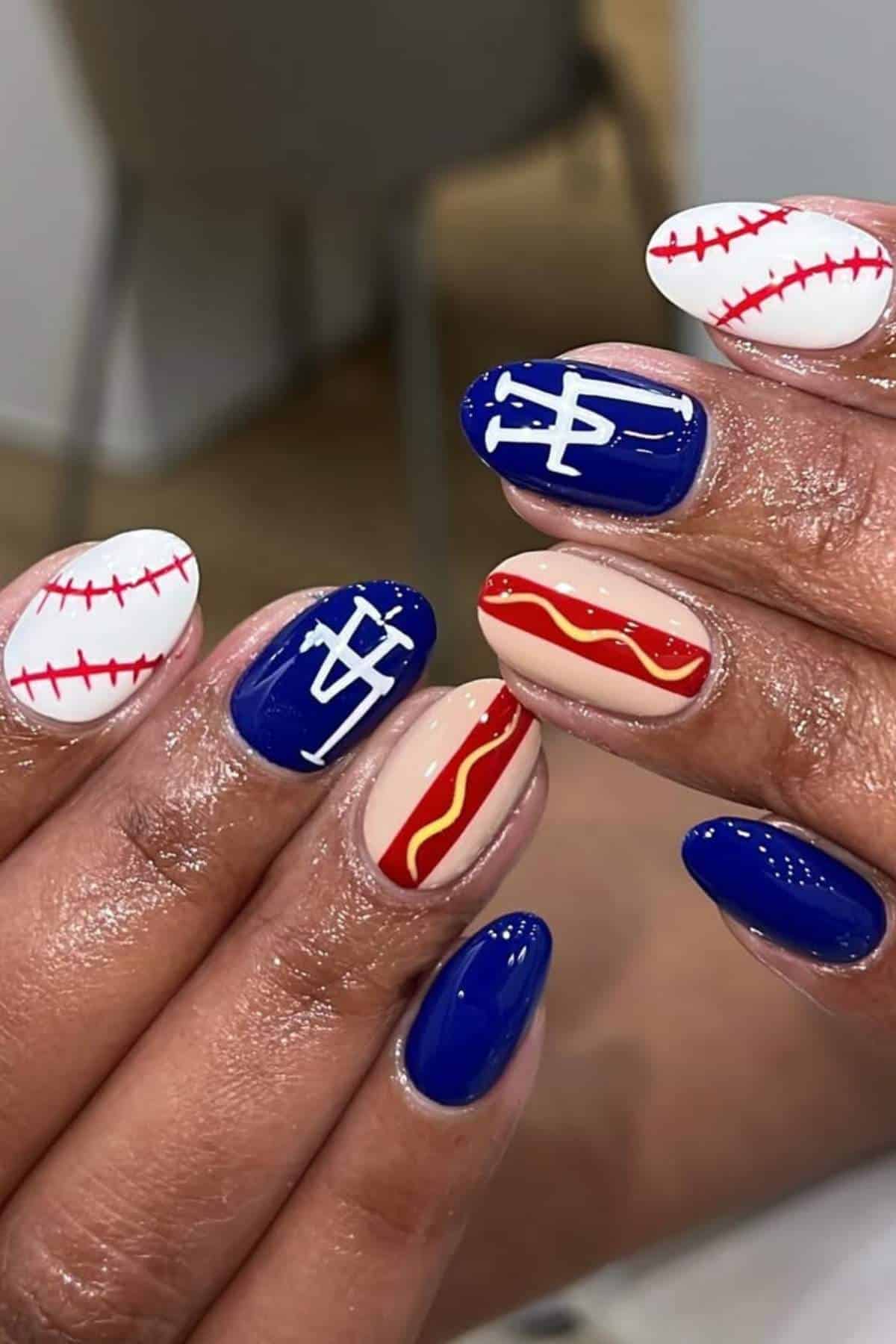 Close-up of hands with baseball-themed nail art, featuring baseball stitching, blue nails with white letters, and a nail decorated with a hot dog design.