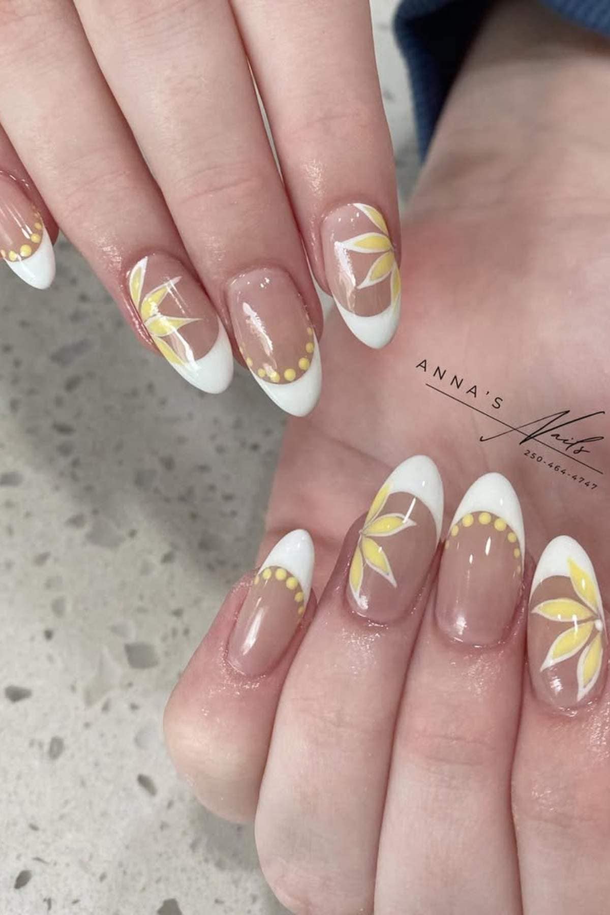 Hands with almond-shaped nails featuring white French tips, yellow dotted accents, and floral designs on a speckled countertop.