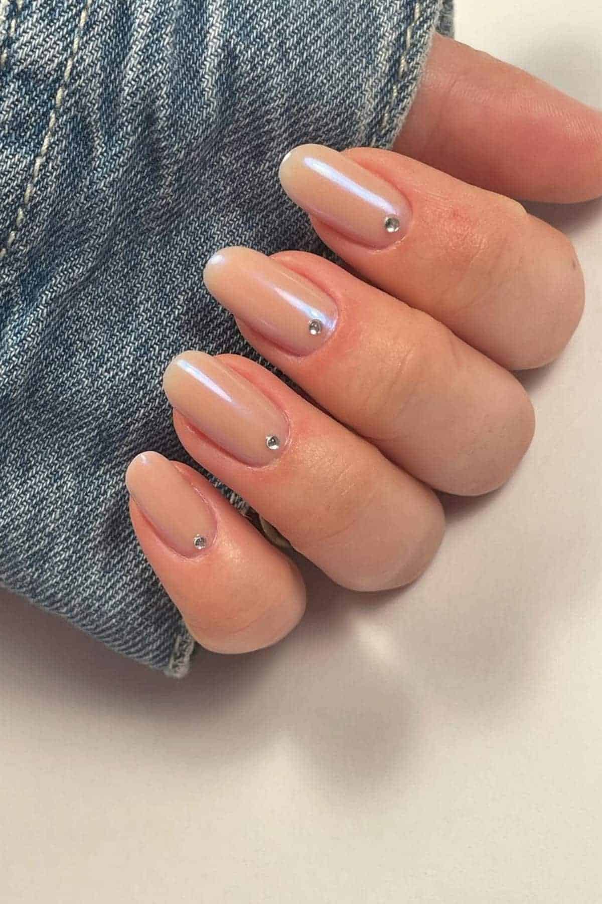 A hand with almond-shaped, iridescent nude nails, each decorated with a small silver stud near the cuticle. The person is wearing a blue denim sleeve.
